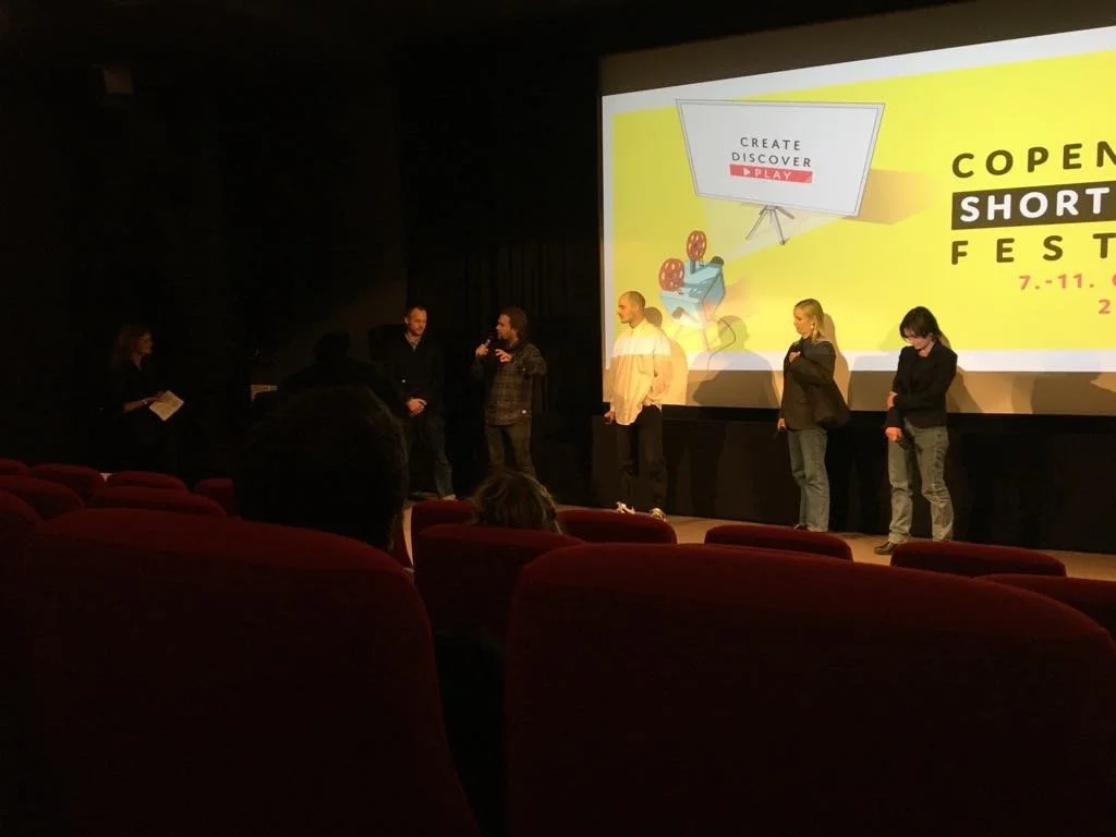 Et hold talere står foran en stor skærm ved Copenhagen Short Film Festival, mens publikum sidder i biografsæder | Kortfilm | In Medias Ræs | Esben Persson | Filmproduktion Aarhus | GESTALT