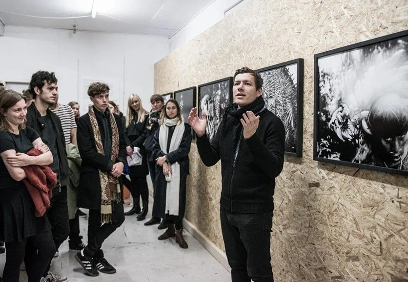 En gruppe mennesker i et fotogalleri. Han gestikulerer med hænderne, mens de andre lytter opmærksomt. mænd holder oplæg | Film- og fotofestival på Aarhus Kunstakademi ved navn Aarhus Independent Pixels | Eventbilleder | GESTALT