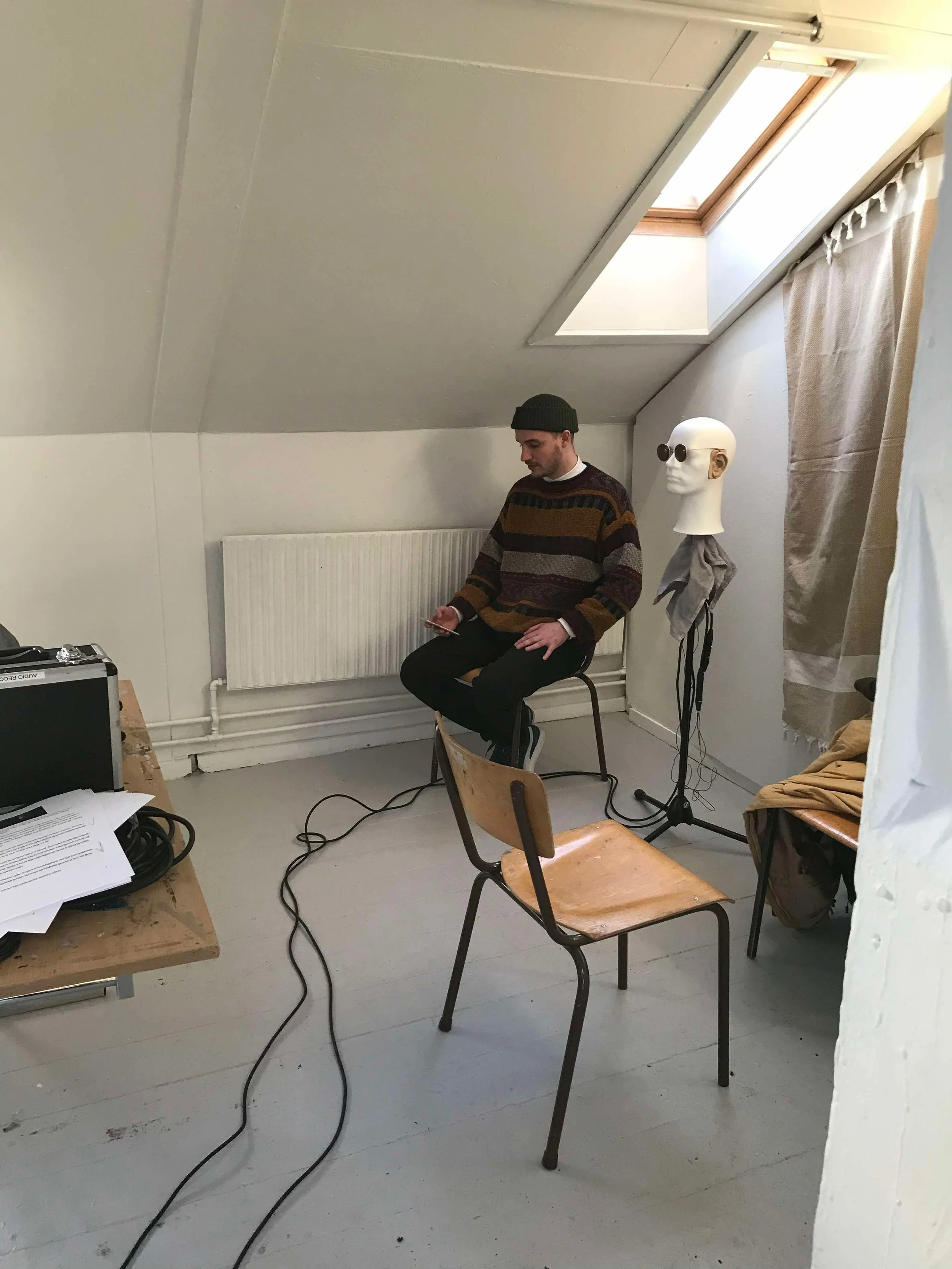 En mand sidder på en stol i et rum med skrå vægge. Han bruger en smartphone. Der står en mannequindukke  ved siden af ham | Musikkomposition | Studieproduktion og lyddesign | Filmproduktion Aarhus | GESTALT