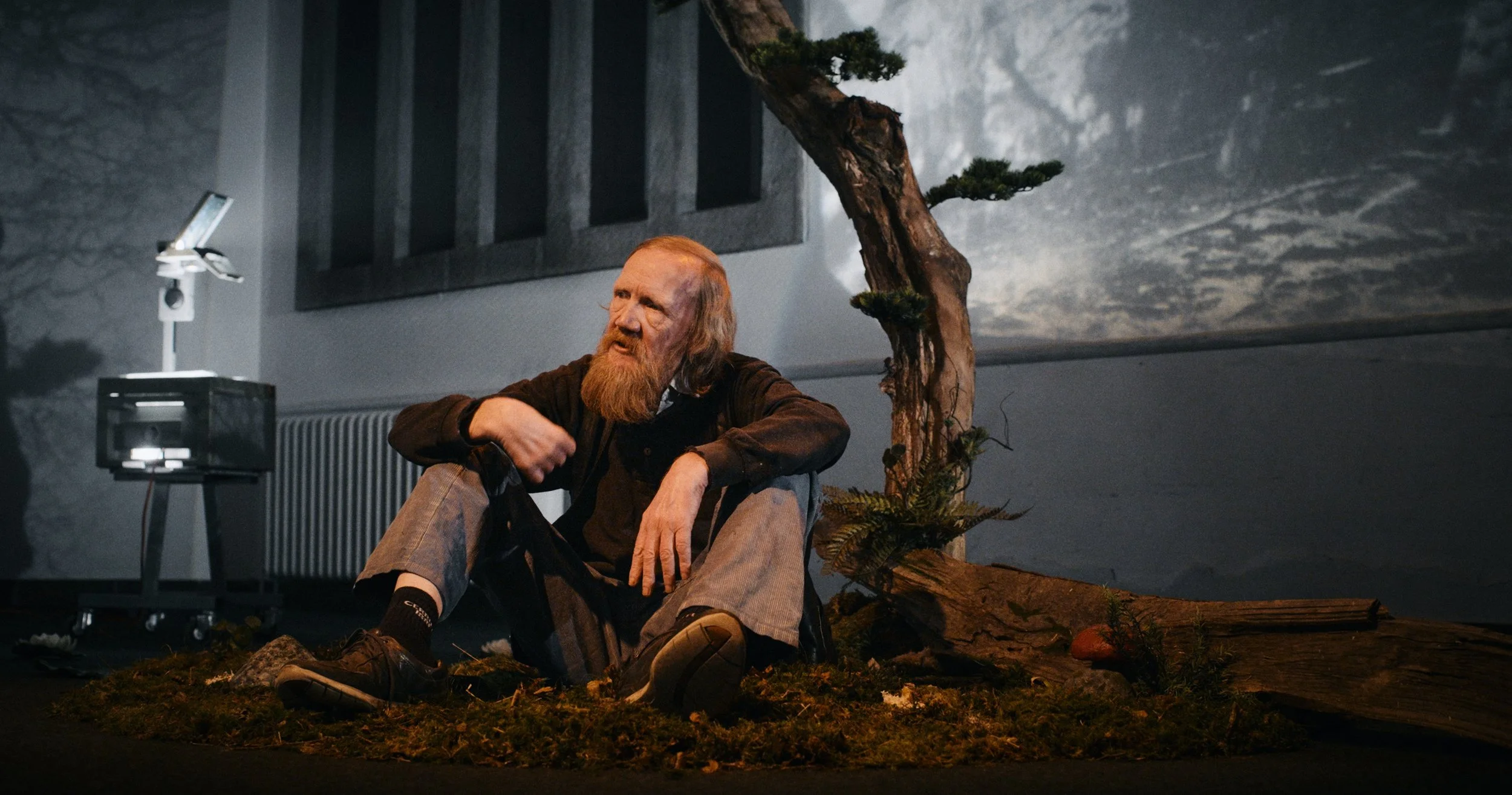En mand med rødt skæg sidder på jorden foran et gammelt træ, omgivet af mos og små planter, i et mørkt indendørs rum | Dokumentarfilm | Retrograd | Esben Persson & Anders Mielcke | Filmproduktion Aarhus | GESTALT