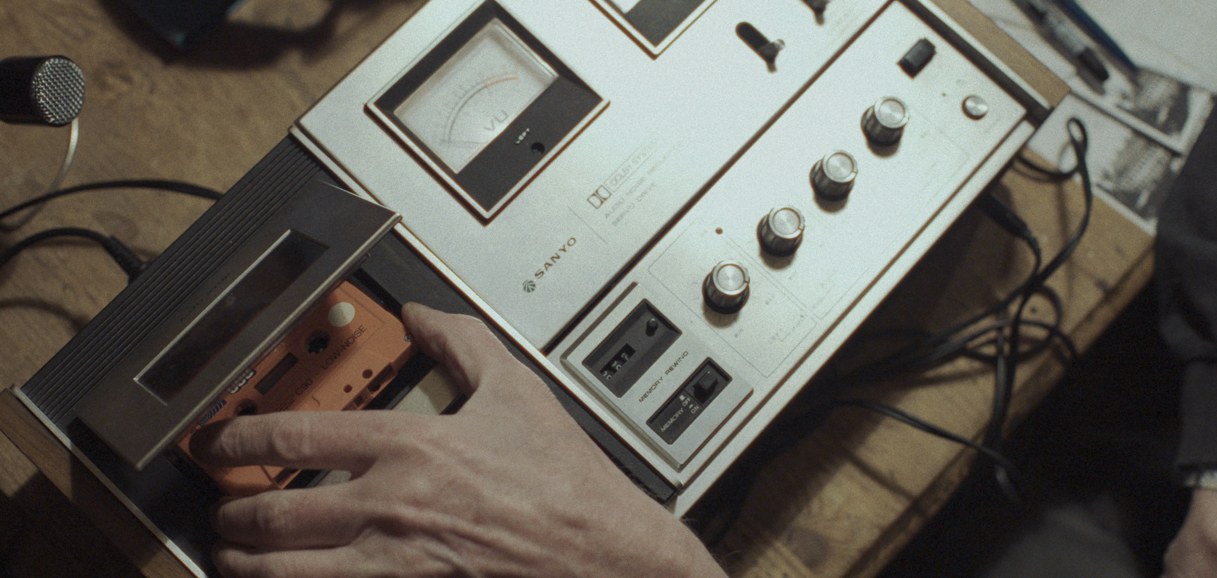 En person bruger en hånd til at håndtere et orange kassettebånd i en båndoptager, placeret på en træbord | Dokumentarfilm | Retrograd | Esben Persson & Anders Mielcke | Filmproduktion Aarhus | GESTALT



