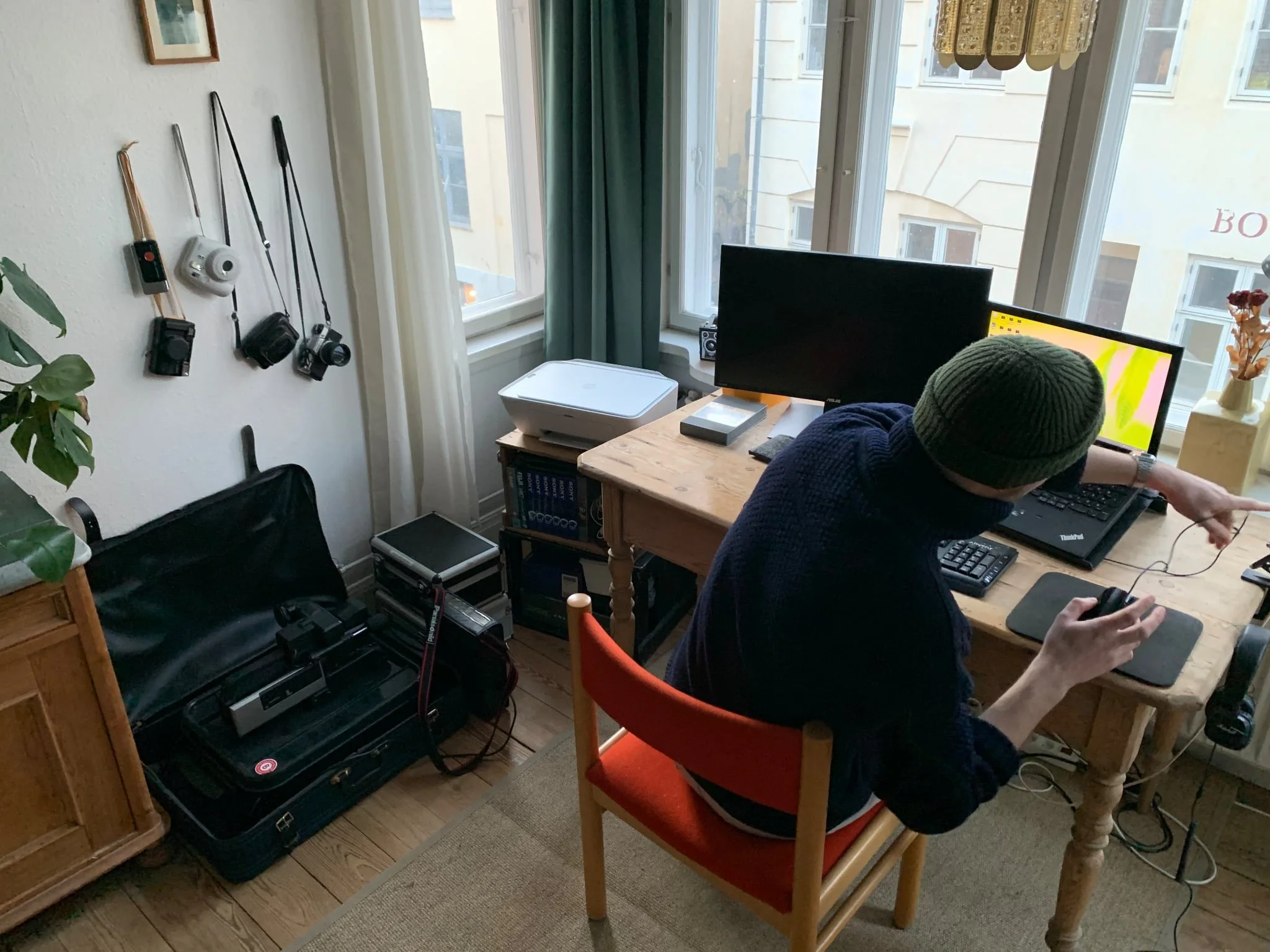 Person med grøn hue, siddende ved skrivebord med to computere og udstyr, i et hjemmekontor med store vinduer, væg med kameraer, og bagage på gulvet | Kortfilm | In Medias Ræs | Bagombilleder | Esben Persson | Filmproduktion Aarhus | GESTALT