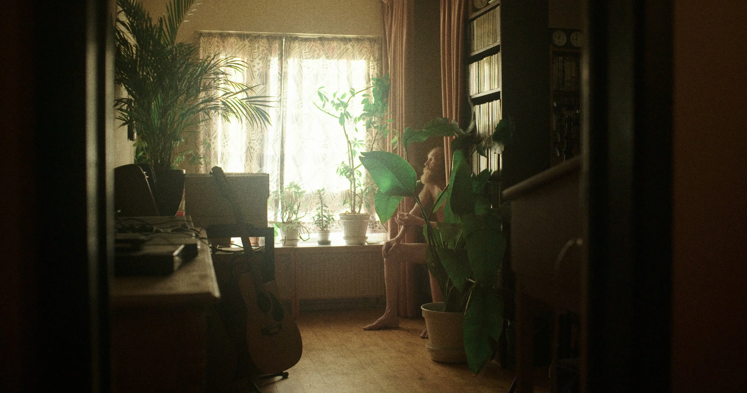 Indendørs scene med et menneske, der sidder i en stol ved et vindue med blomster og planter, og en guitar står på gulvet | Dokumentarfilm | Retrograd | Esben Persson & Anders Mielcke  | Filmproduktion Aarhus | GESTALT