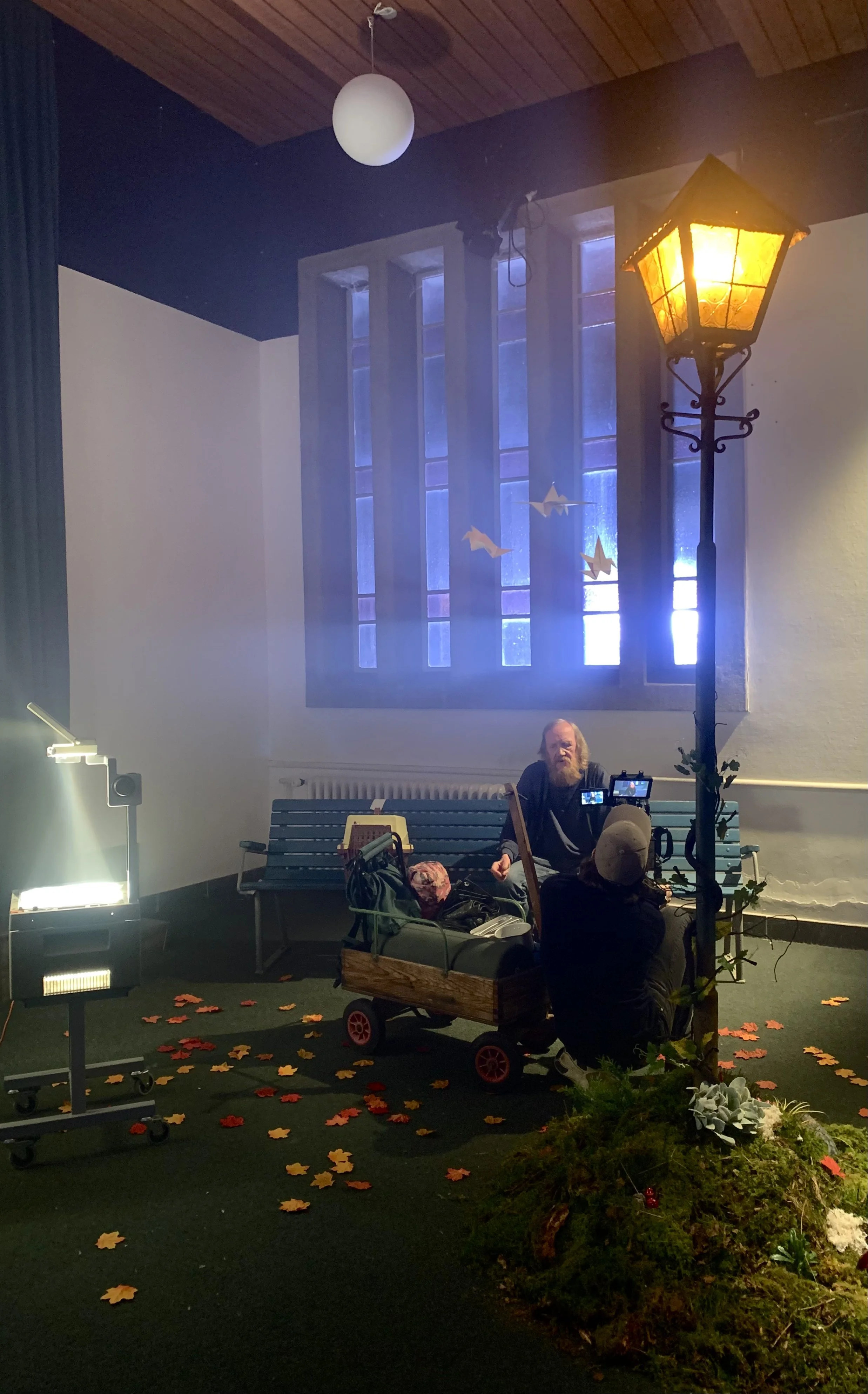 Indendørs scene med en mand, der sidder på en bænk foran et vindue, og bagved en gadelygte, med et filmkamera og en projektor i forgrunden | Dokumentarfilm | Retrograd | Esben Persson & Anders Mielcke | Filmproduktion Aarhus | GESTALT