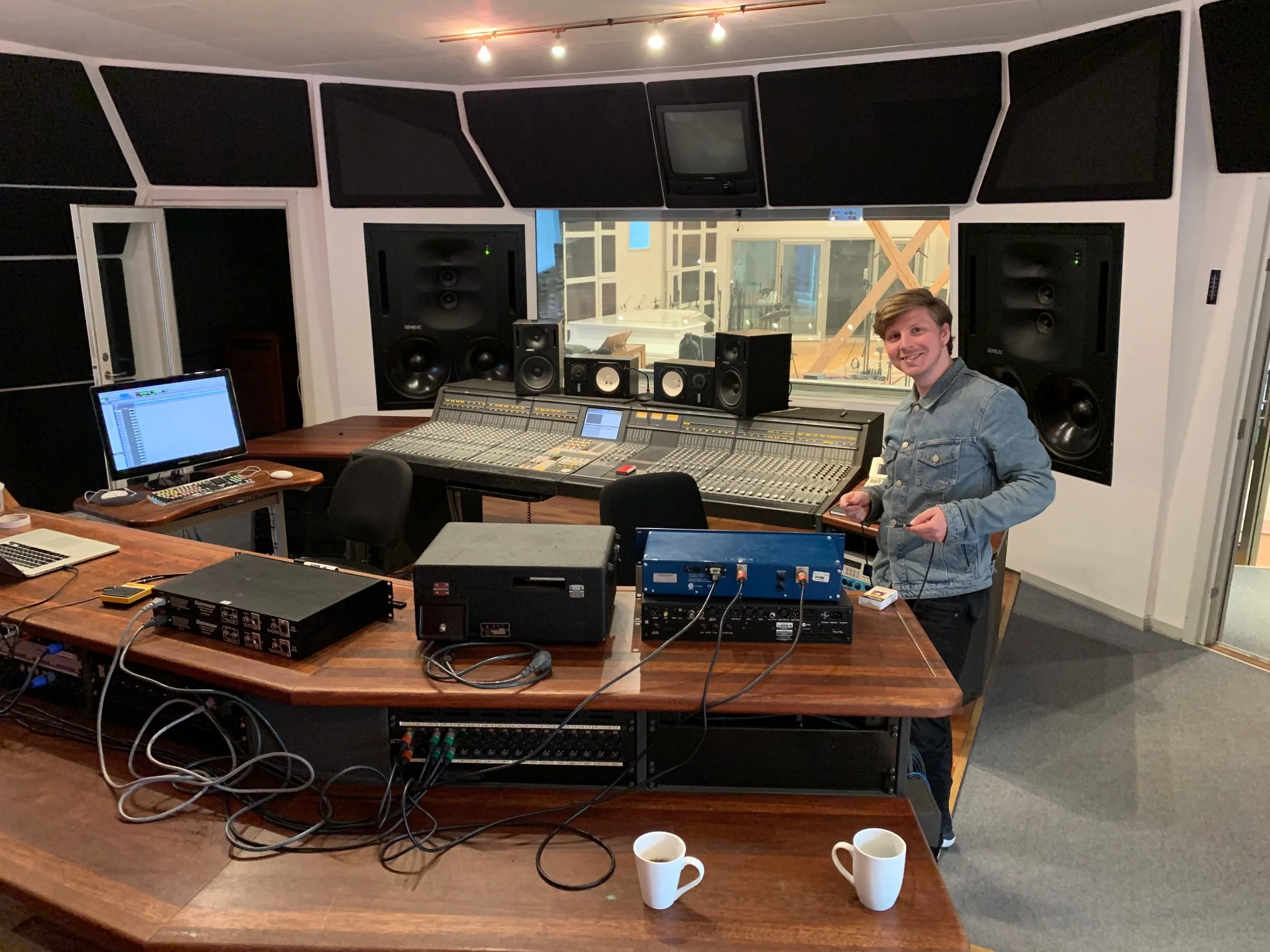 En person står i et lydstudie med stor mixerpult, højtalere, computere og lydudstyr. Personen smiler og holder en ledning, med to kopper kaffe i forgrunden | Musikkomposition | Studieproduktion og lyddesign | Filmproduktion Aarhus | GESTALT