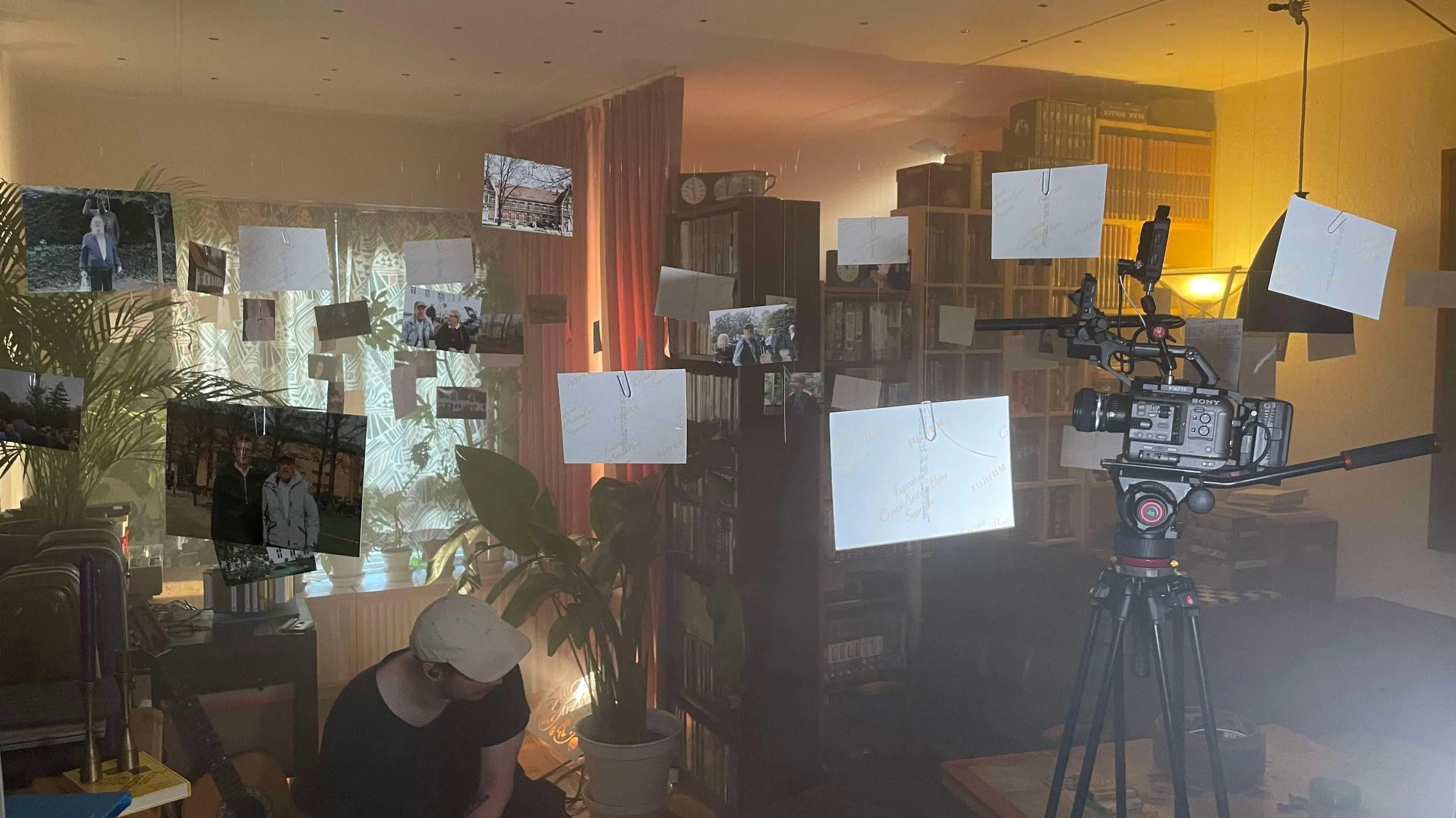 Indendørs scene med to personer og kamera, omgivet af fotos hængt op i et rum med bogreoler og planter | Dokumentarfilm | Retrograd | Esben Persson & Anders Mielcke | Filmproduktion Aarhus | GESTALT