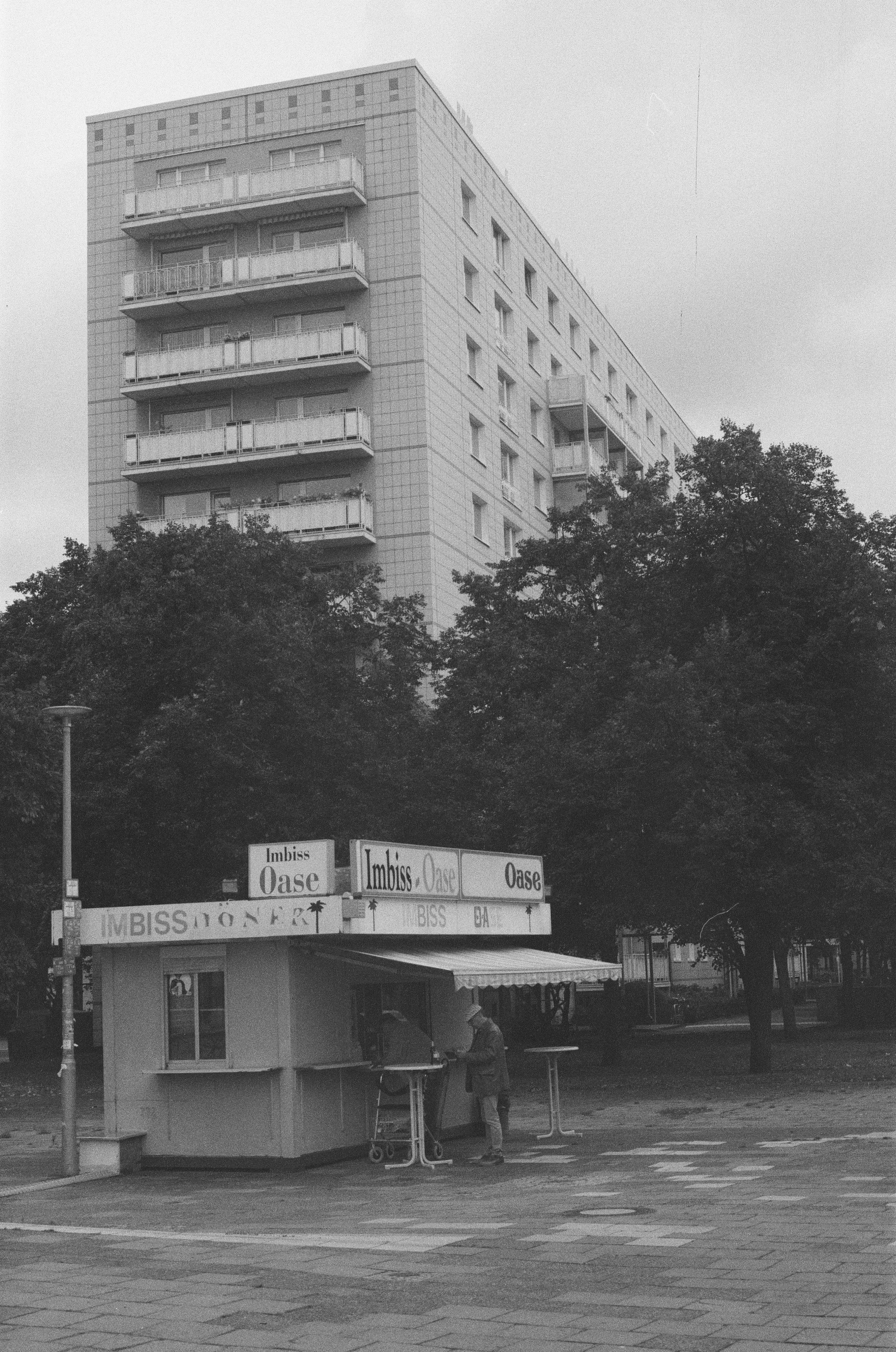 Et enkelt byggeri med en høj boligblok bag en kiosk, der sælger snacks og sodavand, og der står to personer foran, i en urban dagligdagsscene | Location scouting | Kodak | Analoge optagelser | Filmproduktion Aarhus | GESTALT
