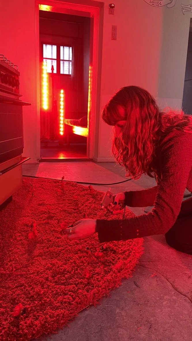 En kvinde med glat hår, der sidder på gulvet og bruger et værktøj på en rød tæppe, i et rum med en dør, der er oplyst af rødt lys fra en lysstofarmatur | Dokumentarfilm | Retrograd | Esben Persson & Anders Mielcke  | Filmproduktion Aarhus | GESTALT