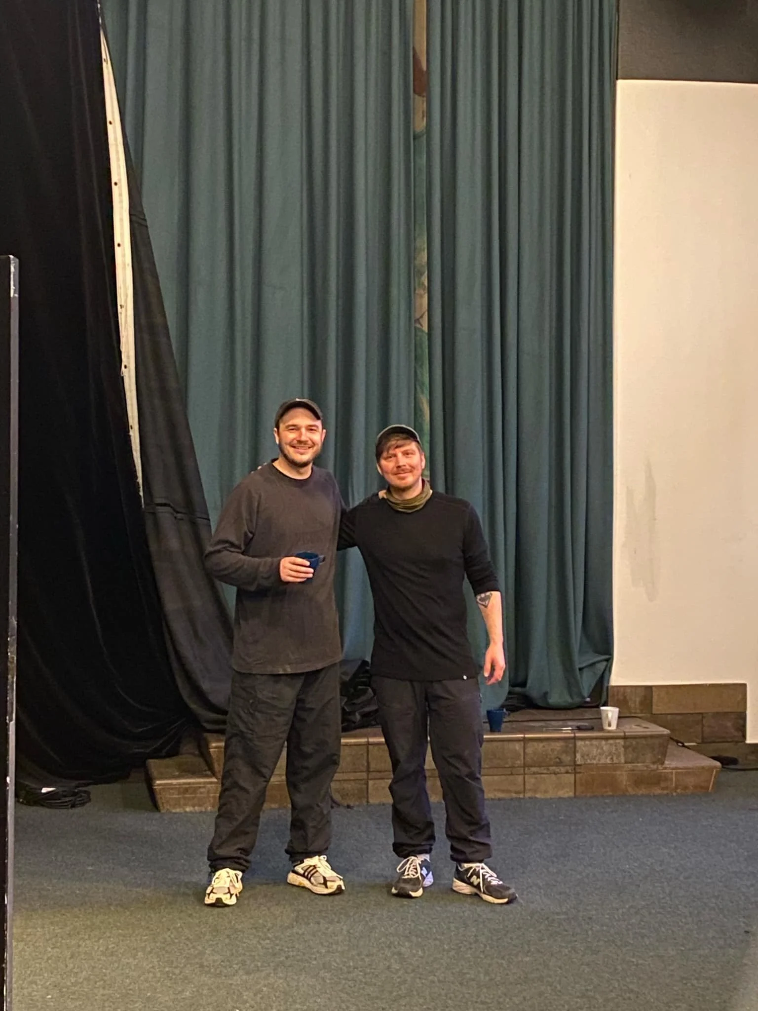 To mænd står side om side, smilende, foran en grøn scenetæppe og en sort scenedraperie, på et gulv med tæppe | Dokumentarfilm | Retrograd | Esben Persson & Anders Mielcke | Filmproduktion Aarhus | GESTALT