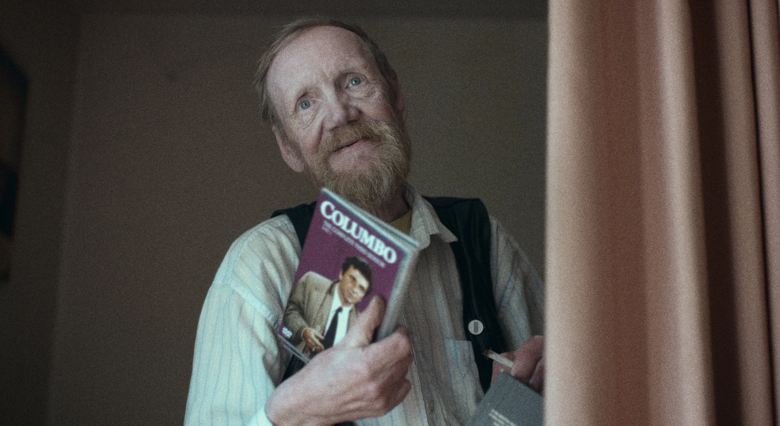 En ældre mand med rødbrunt skæg og lyst hår, der holder et blad med en tegneserie af en mand i jakkesæt, foran en lyserød væg og en delvist trukket brun gardin | Dokumentarfilm | Retrograd | Esben Persson & Anders Mielcke  | Filmproduktion Aarhus 