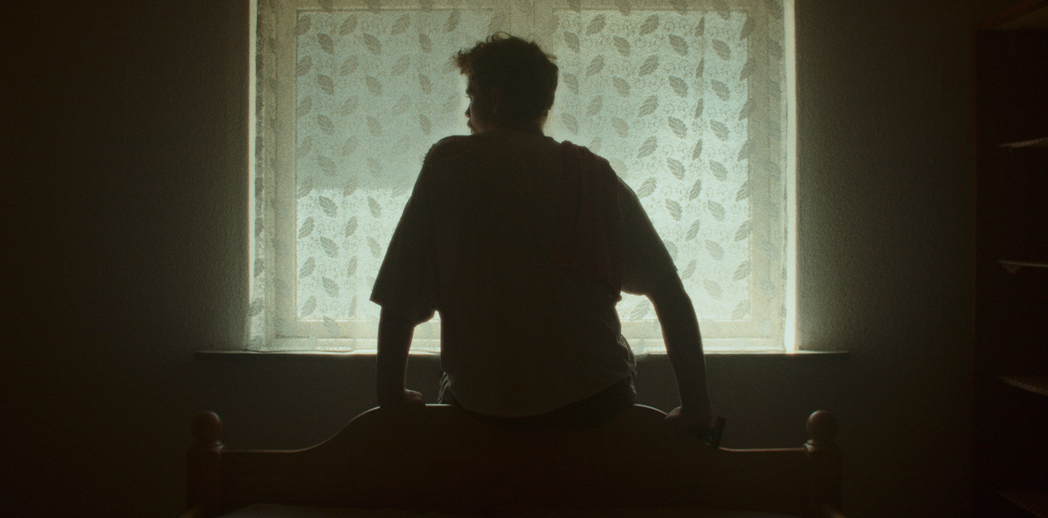 Silhuet af en person, der sidder på en seng foran et vindue med gardin, i et dunkelt værelse | Kortfilm | Wild Wild Vesløs | Esben Persson | Filmproduktion Aarhus | GESTALT