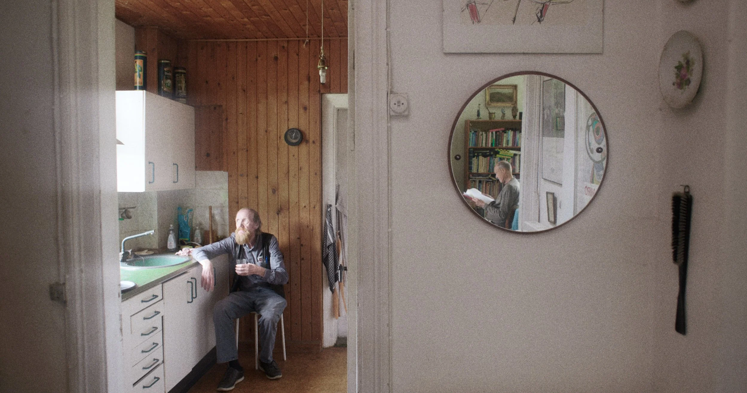 En mand med skæg sidder i et køkken og holder et glas. Der er et rundt spejl på væggen, der viser en anden person, der læser en bog. Køkkenet har hvide skabe og en grøn vask | Dokumentarfilm | Retrograd | Esben Persson & Anders Mielcke  | GESTALT