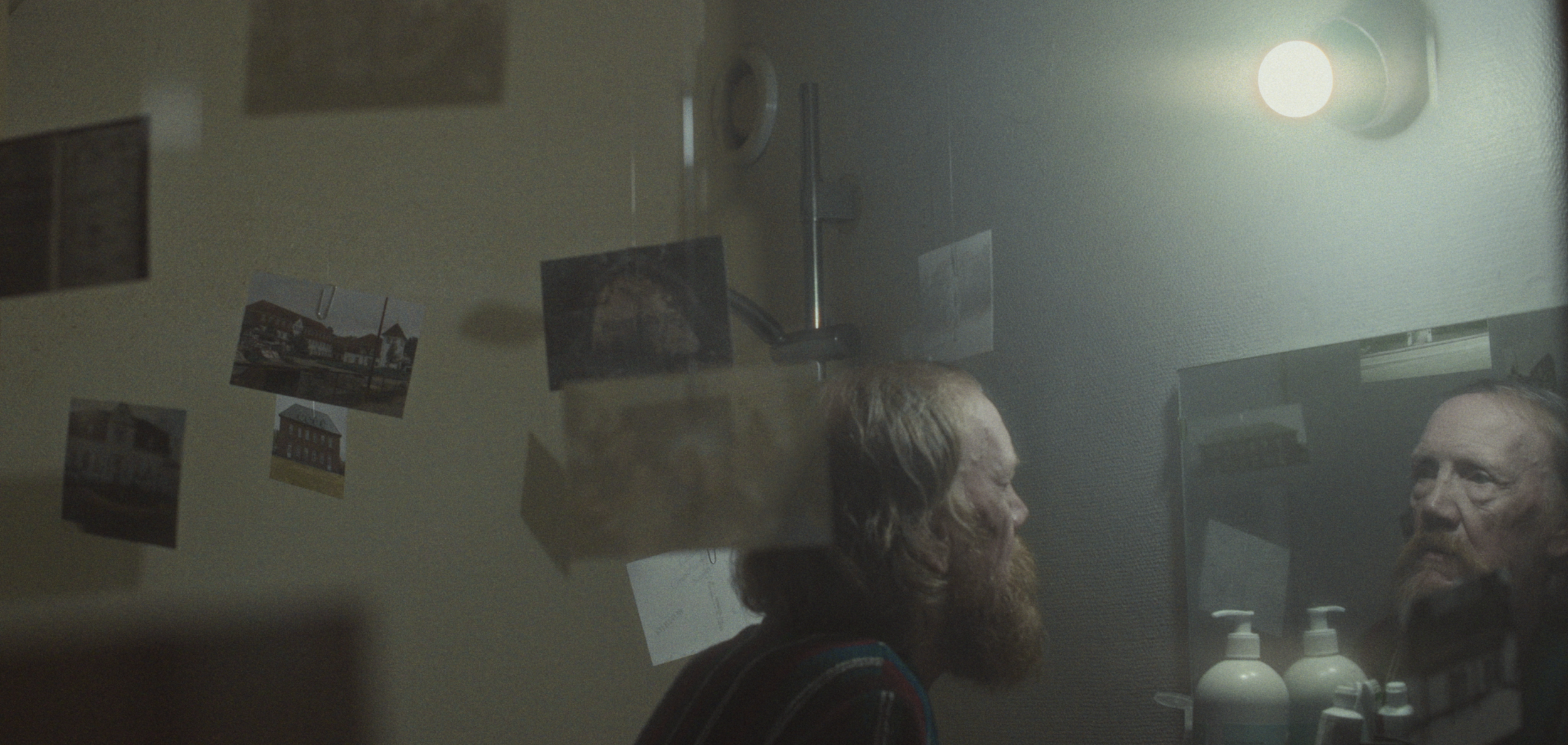 En mand med skæg ser sig selv i spejlet på sit badeværelse, med fotos hængende ned fra loftet | Dokumentarfilm | Retrograd | Esben Persson & Anders Mielcke | Filmproduktion Aarhus | GESTALT