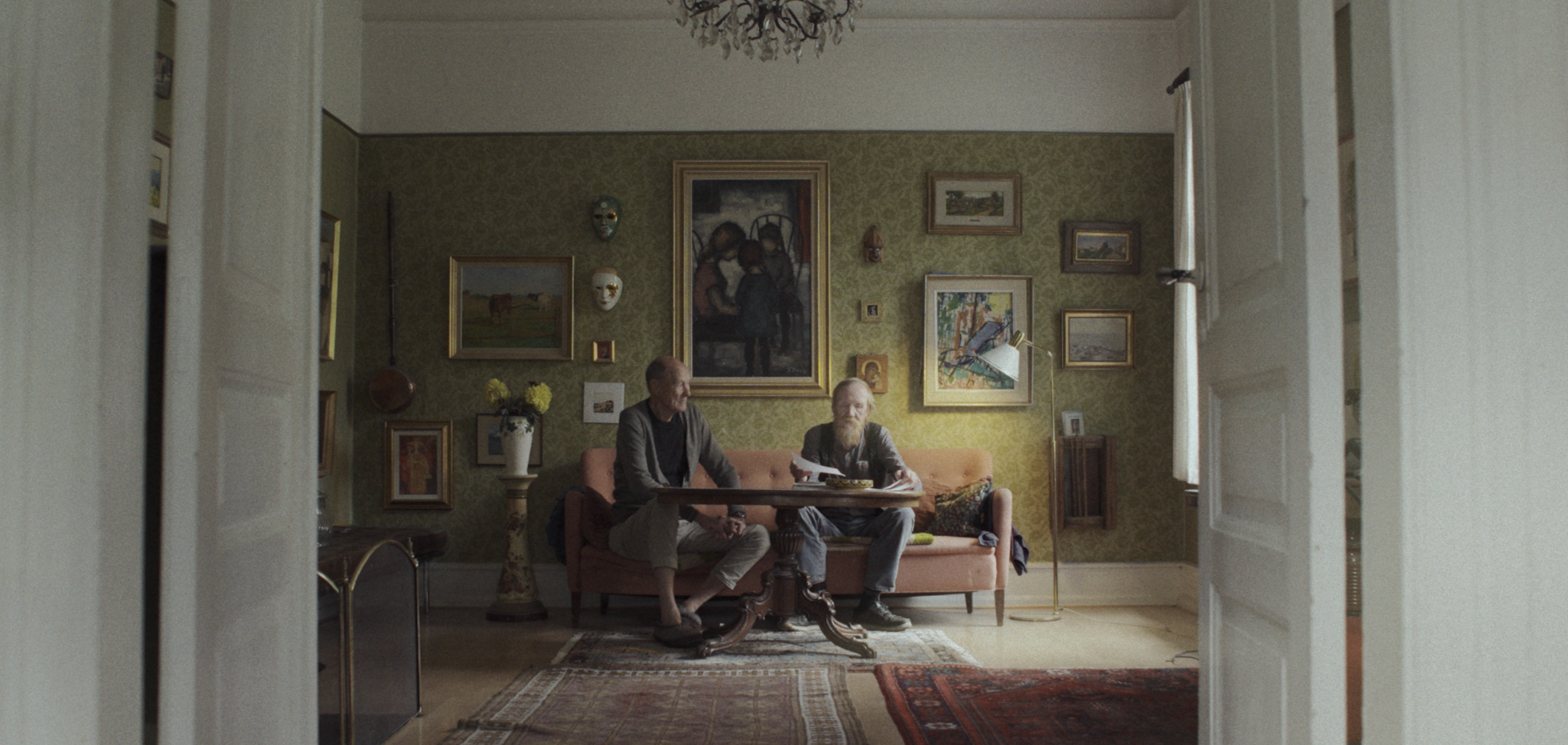 To mænd sidder på en rosa sofa i en stue, der er pyntet med mange kunstværker og masker på væggen | Dokumentarfilm | Retrograd | Esben Persson & Anders Mielcke | Filmproduktion Aarhus | GESTALT