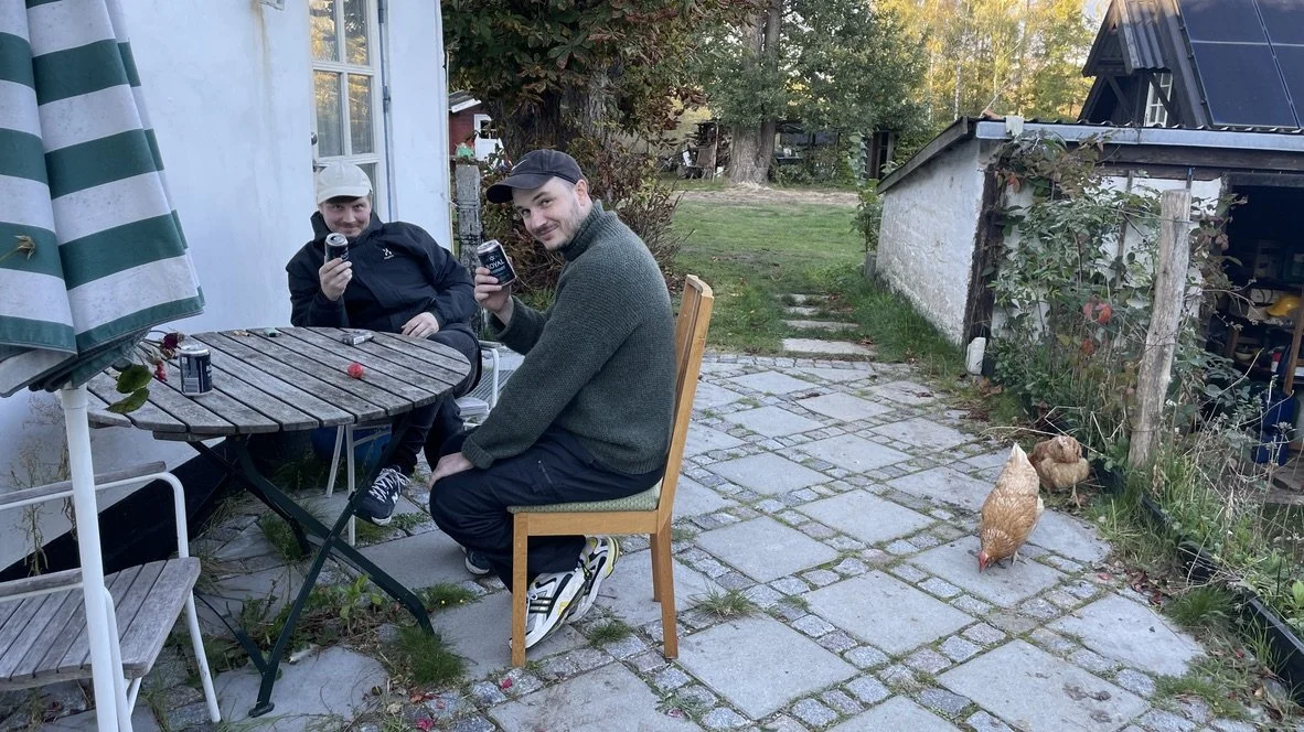 To mænd sidder ude på en terrasse, nyder drikkevarer og smiler til kameraet. Der er en stol og et bord, et par høns, en parasol, og en have med grønne træer i baggrunden | Dokumentarfilm | Retrograd | Esben Persson & Anders Mielcke | Filmproduktion 