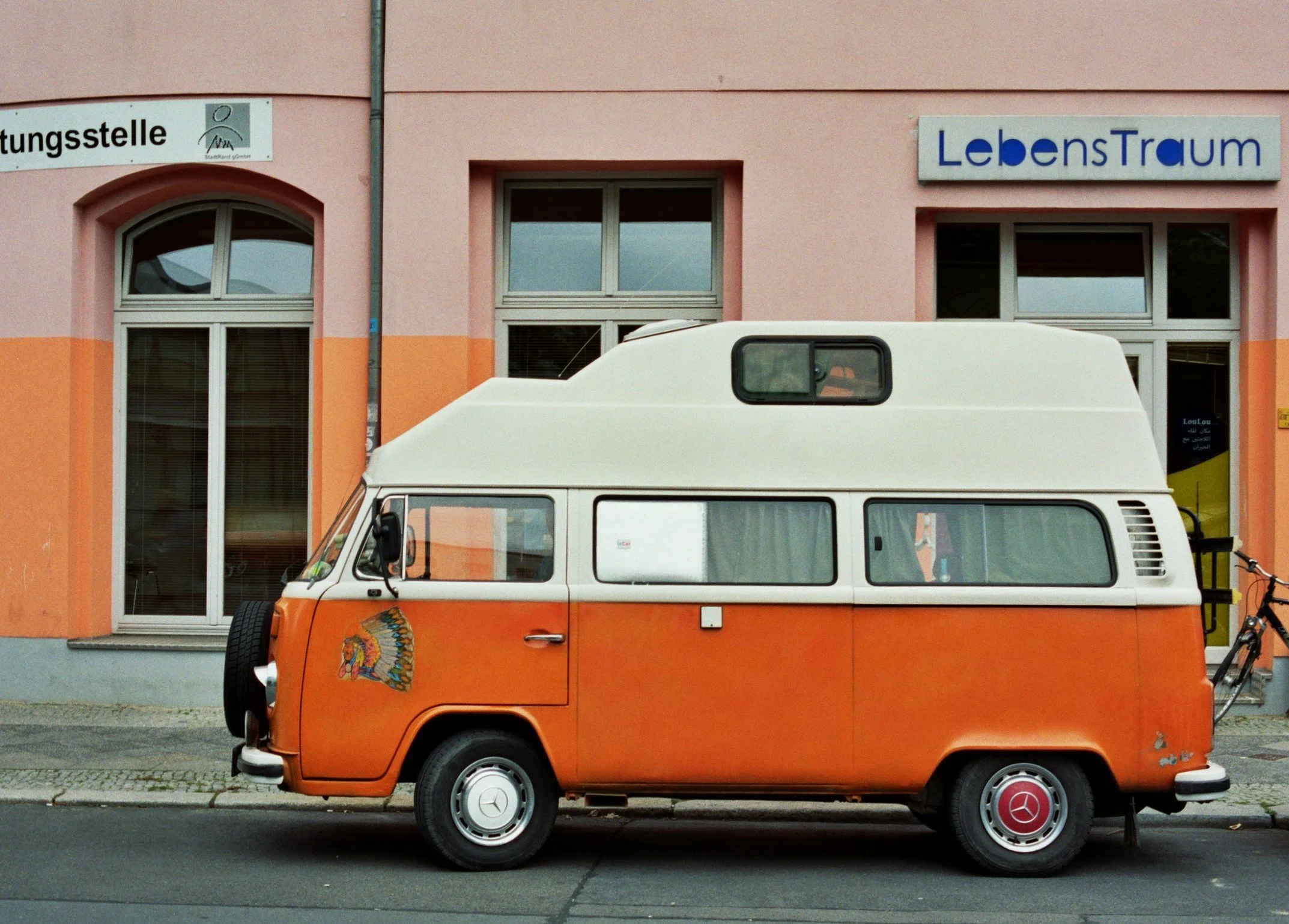 En vintage orange og hvid campingvogn parkeret foran en bygning med blomstrende vinduer, en cykel fastgjort til bagenden | Location scouting Berlin | Kodak | Filmproduktion Aarhus | GESTALT