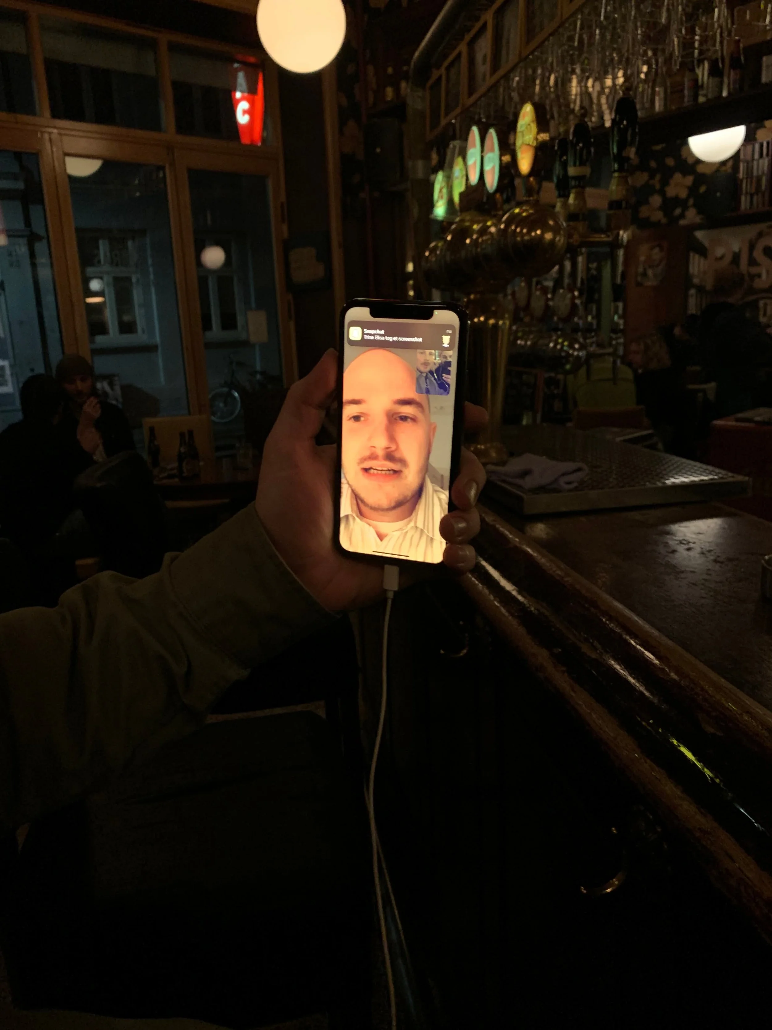 Person viser en telefon med et ansigtsvideoopkald til en anden person i en hyggelig bar eller café med tavler og lamper.
