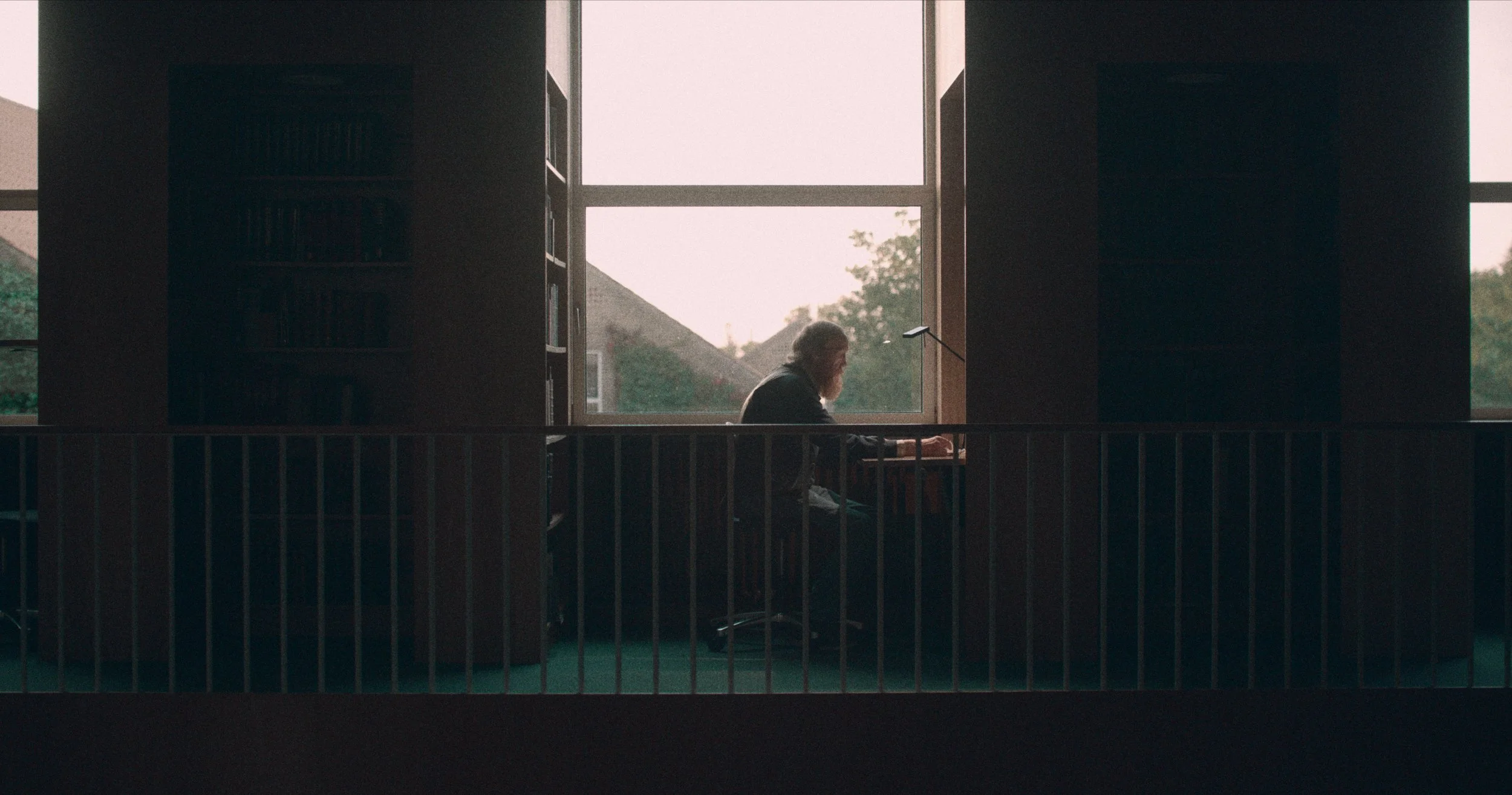 En person med langt hår sidder ved et skrivebord foran et vindue, der viser huse og træer udenfor, i en rolig indendørs indretning | Dokumentarfilm | Retrograd | Esben Persson & Anders Mielcke | Filmproduktion Aarhus | GESTALT