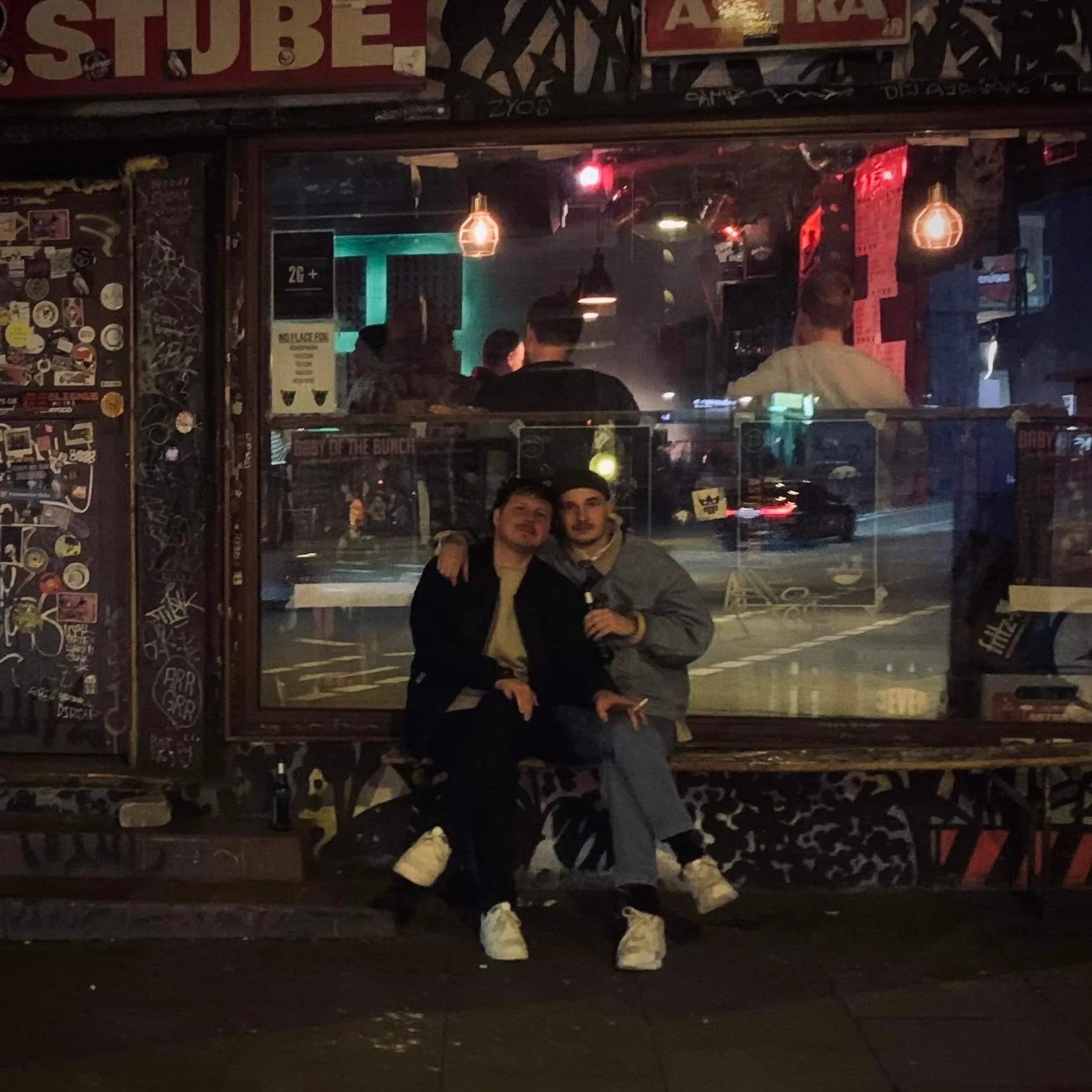 To unge mænd sidder på en bænk foran en bar, hvor flere mennesker spiser og drikker indendørs. Billedet er taget om aftenen, og der er dekorative graffiti og stickers på væggene omkring dem | Stemningsbillede | Filmproduktion i Aarhus | GESTALT