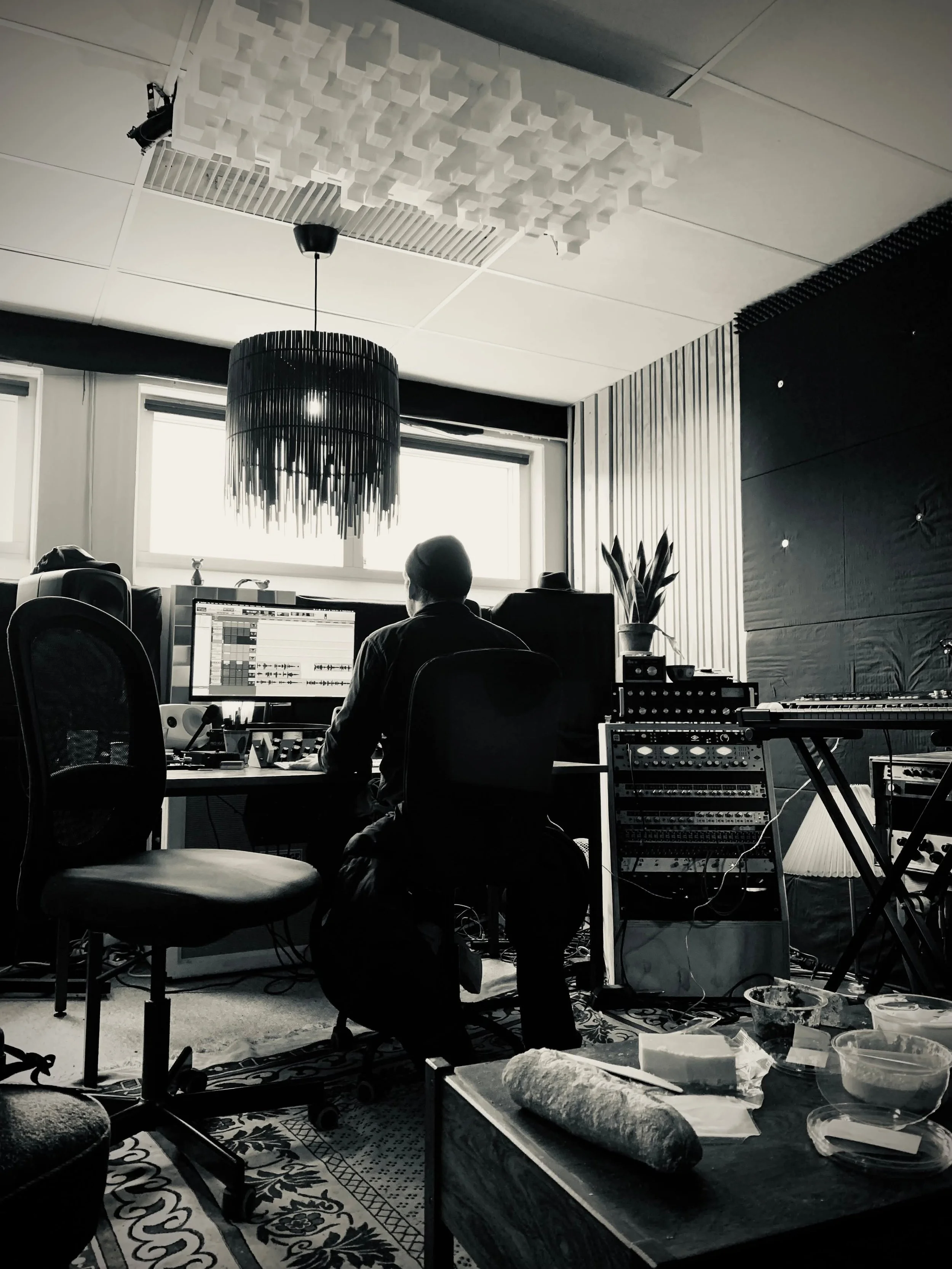 Person der arbejder i et musikstudie med computer og lydudstyr, omgivet af musikinstrumenter og optagelsesudstyr | Studieproduktion | Musikkomposition | Filmproduktion Aarhus | GESTALT