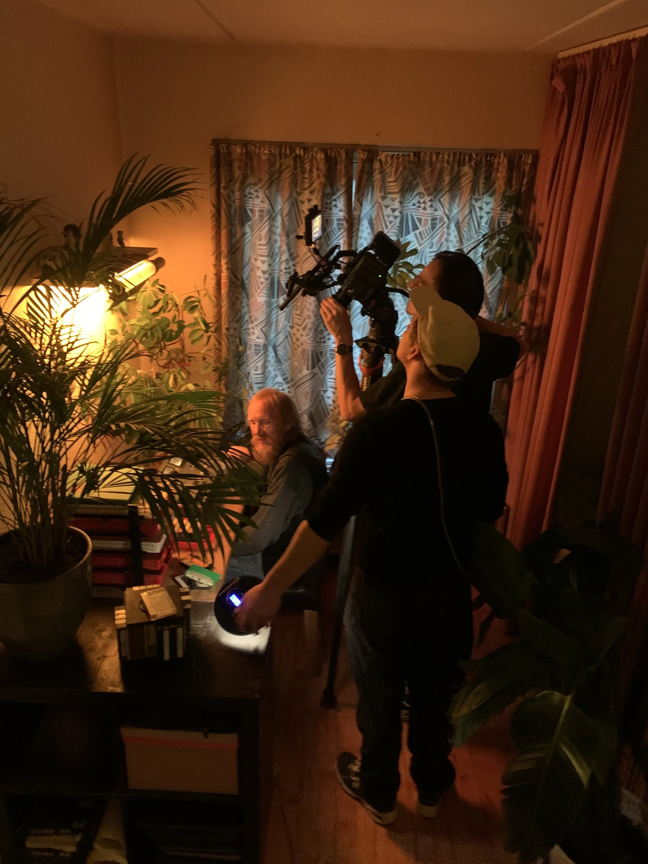 Filminstruktør og fotograf, der optager en samtale med en mand, der sidder ved et bord i et værelse med planter og tæpper | Dokumentarfilm | Retrograd | Esben Persson & Anders Mielcke | Filmproduktion Aarhus | GESTALT