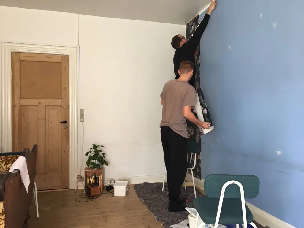 To personer, der monterer eller justerer tapet på en væg i et rum, hvor den ene person står på en stol og den anden hjælper | Kortfilm | In Medias Ræs | Bagombilleder | Esben Persson | Filmproduktion Aarhus | GESTALT