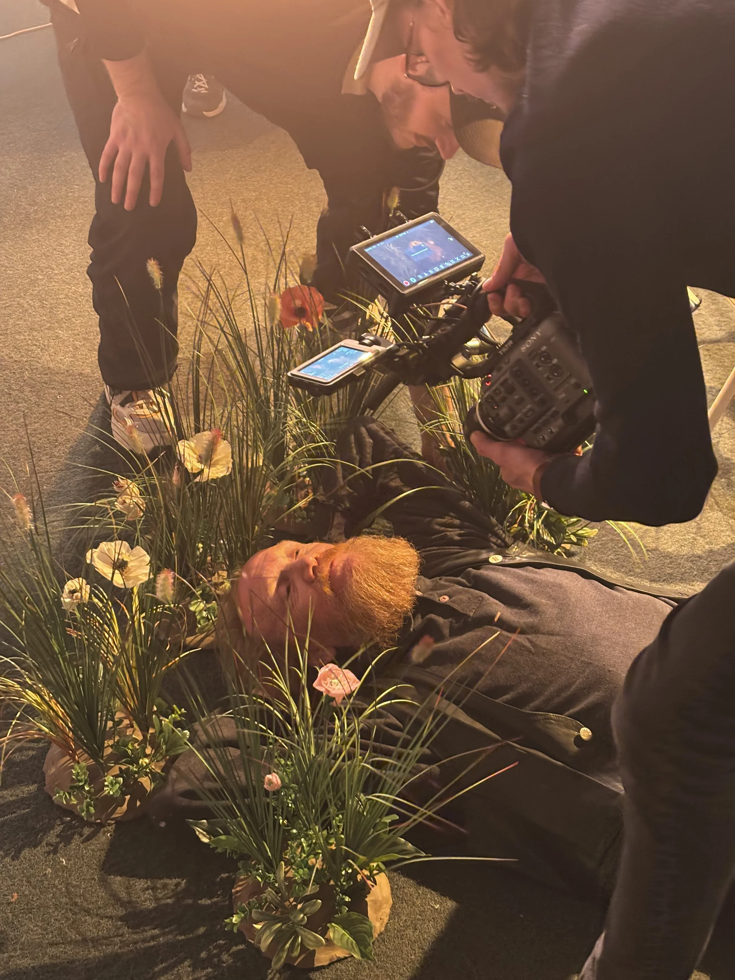 Person ligger på jorden omgivet af blomster og planter, mens andre personer er omkring og filmer ham | Dokumentarfilm | Retrograd | Esben Persson & Anders Mielcke | Filmproduktion Aarhus | GESTALT