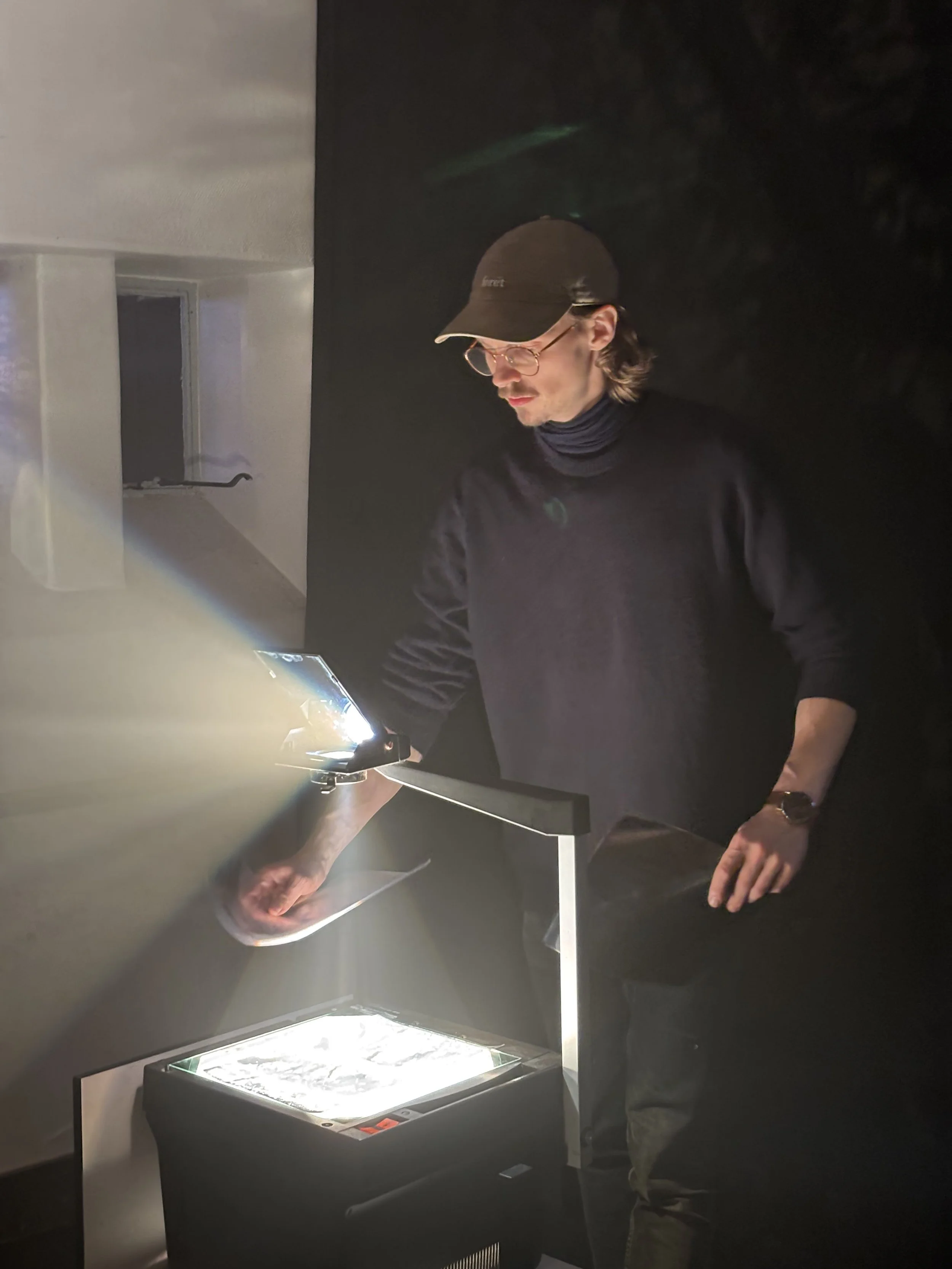 En mand med brunt hår, briller, sort hat og sort turtleneck arbejder med lyssætning eller et udstyr i et mørkt filmstudie | Dokumentarfilm | Retrograd | Esben Persson & Anders Mielcke | Filmproduktion Aarhus | GESTALT.