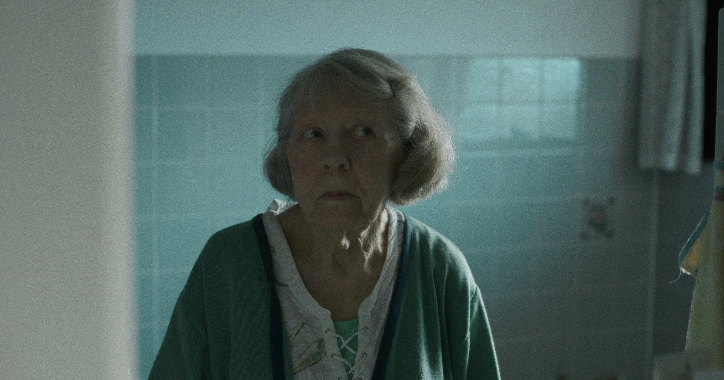En ældre kvinde med gråt hår, der står i et badværelse, iført en grøn cardigan og en hvid bluse, ser på sig selv i spejlet | Dokumentarfilm | Retrograd | Esben Persson & Anders Mielcke | Filmproduktion Aarhus | GESTALT