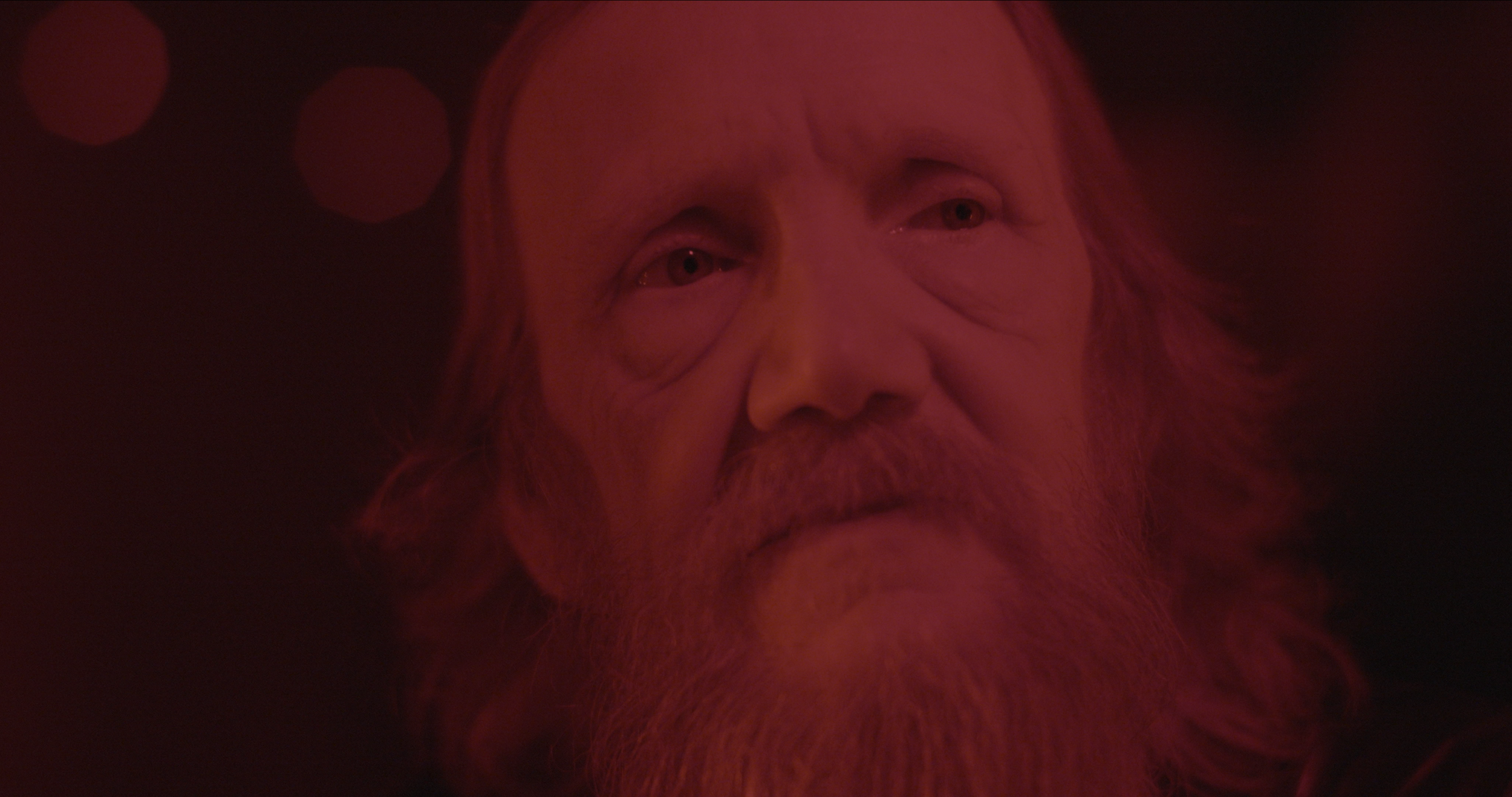 Nærbillede af en ældre mand med langt, gråt hår og skæg, belyst med rødt lys, med en seriøs udtryksfuld mine | Dokumentarfilm | Retrograd | Esben Persson & Anders Mielcke | Filmproduktion Aarhus | GESTALT