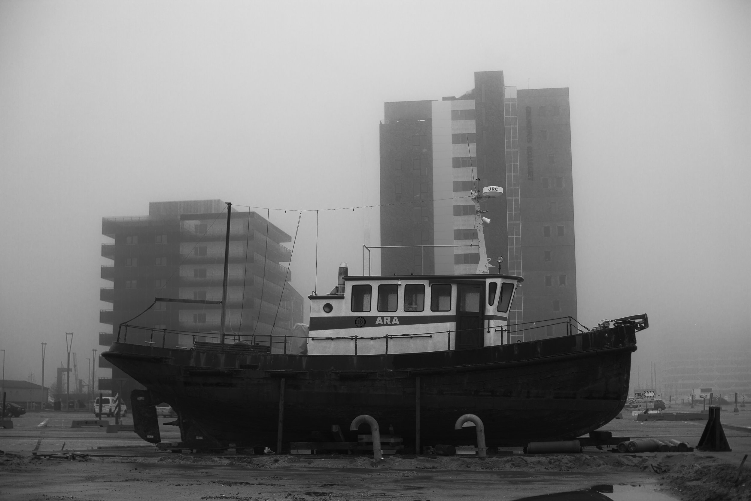En gammel redningsbåd med navnet 'ARA' står på en dock ved en stormfuld og tåget by, omgivet af moderne bygninger i baggrunden | Location scouting | Kodak | Filmproduktion Aarhus | GESTALT