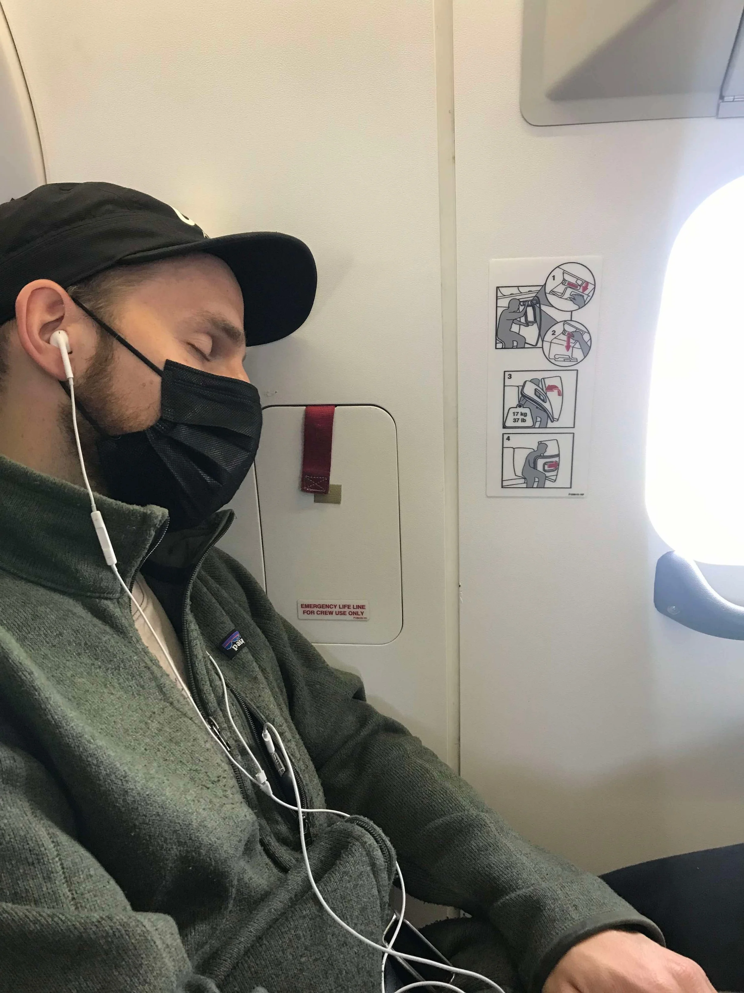 En mand sover siddende med øjne lukkede, iført ansigtsmaske, en sort hat, og øretelefoner, siddende i et flysæde ved vinduet.