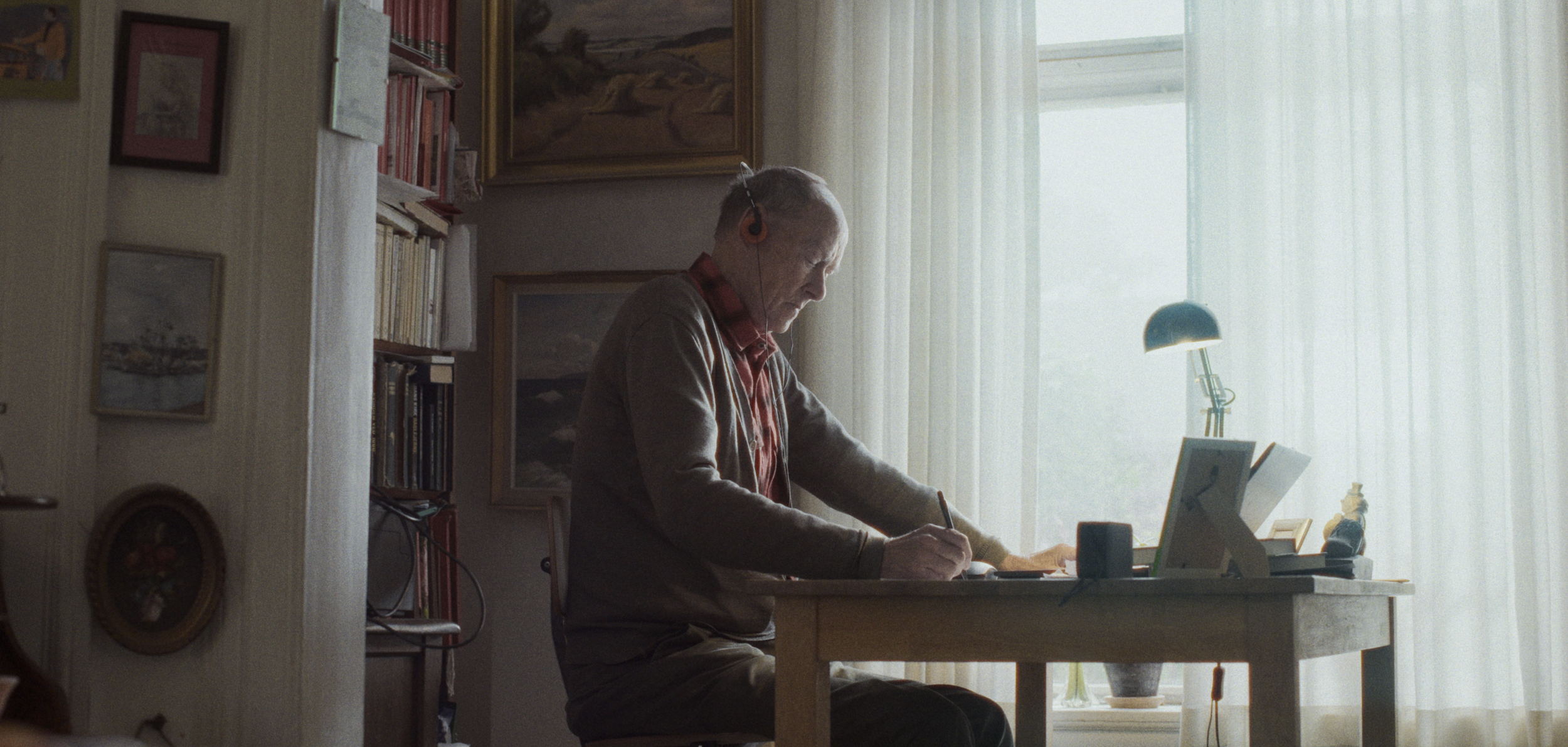 En ældre mand sidder ved et skrivebord, skriver, og har høretelefoner på, i et lyst værelse med bogreoler og kunst på væggen | Dokumentarfilm | Retrograd | Esben Persson & Anders Mielcke | Filmproduktion Aarhus | GESTALT