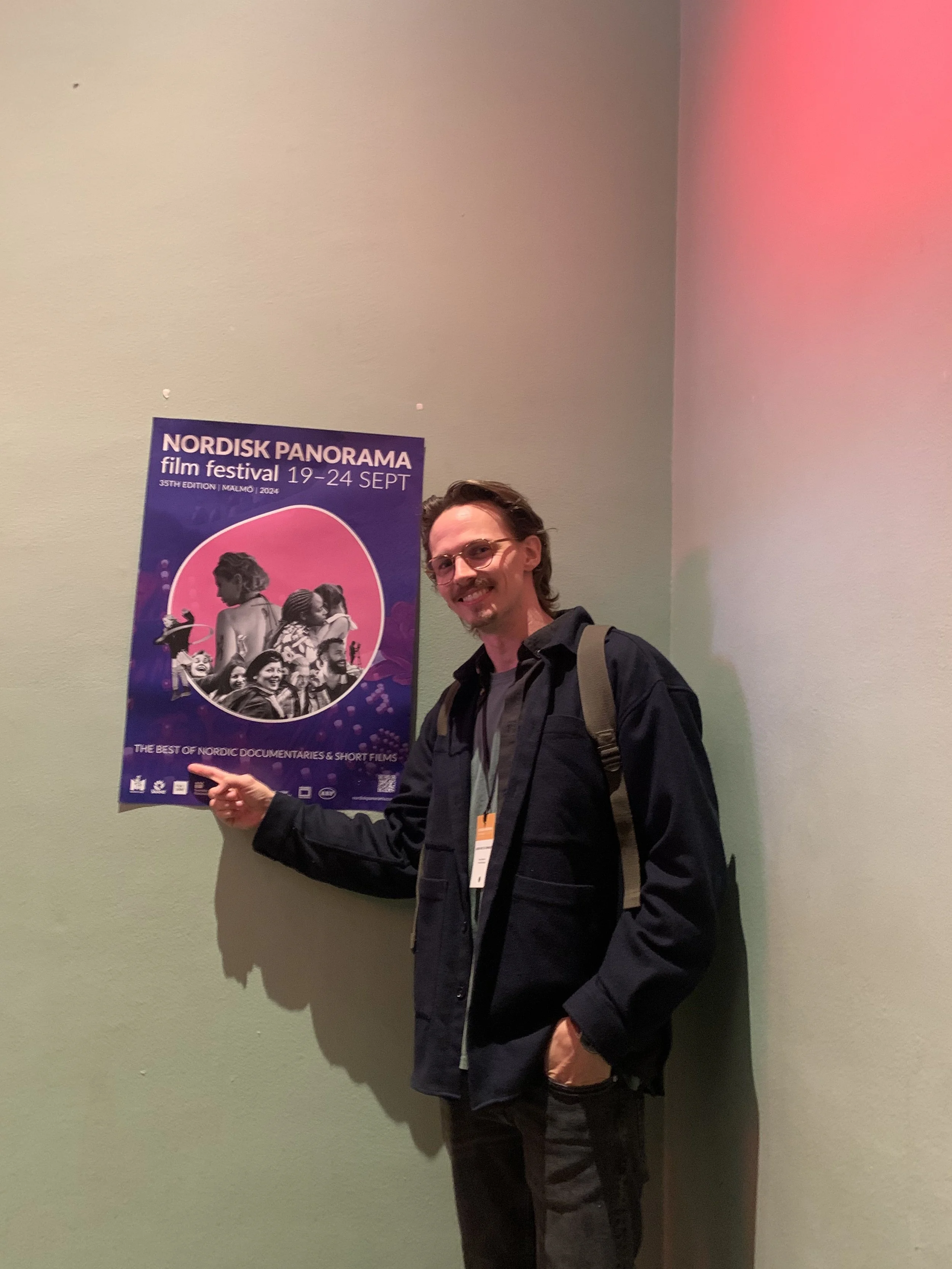En mand, der står foran en plakat for Nordisk Panorama filmfestival, 19-24 september, og peger på den med et smil | Dokumentarfilm | Retrograd | Esben Persson & Anders Mielcke | Filmproduktion Aarhus | GESTALT