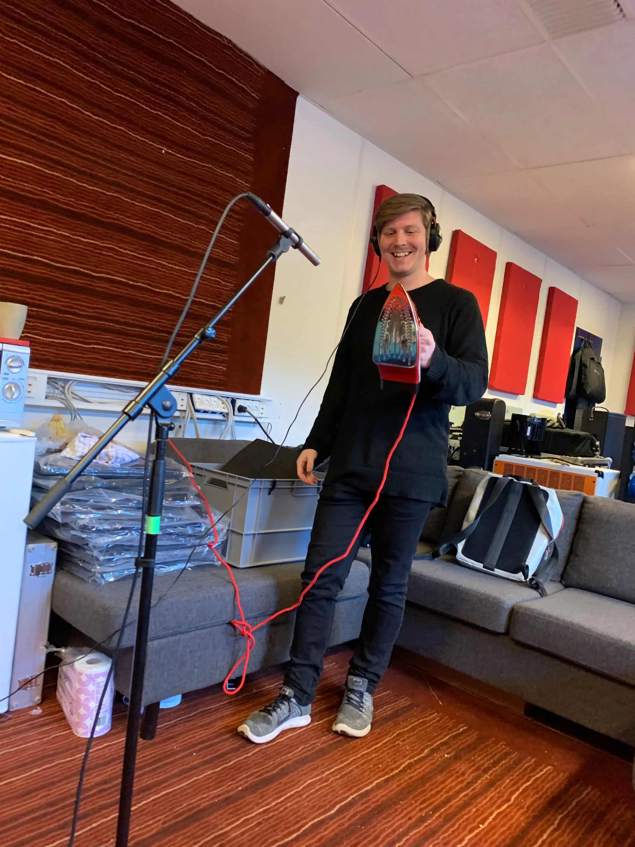 En glad mand står i et lydstudie og holder en guitar med elektriske ledninger, mens han har hovedtelefoner på. Der er mikrofon og lydudstyr omkring ham | Studieproduktion | Musikkomposition | Filmproduktion Aarhus | GESTALT