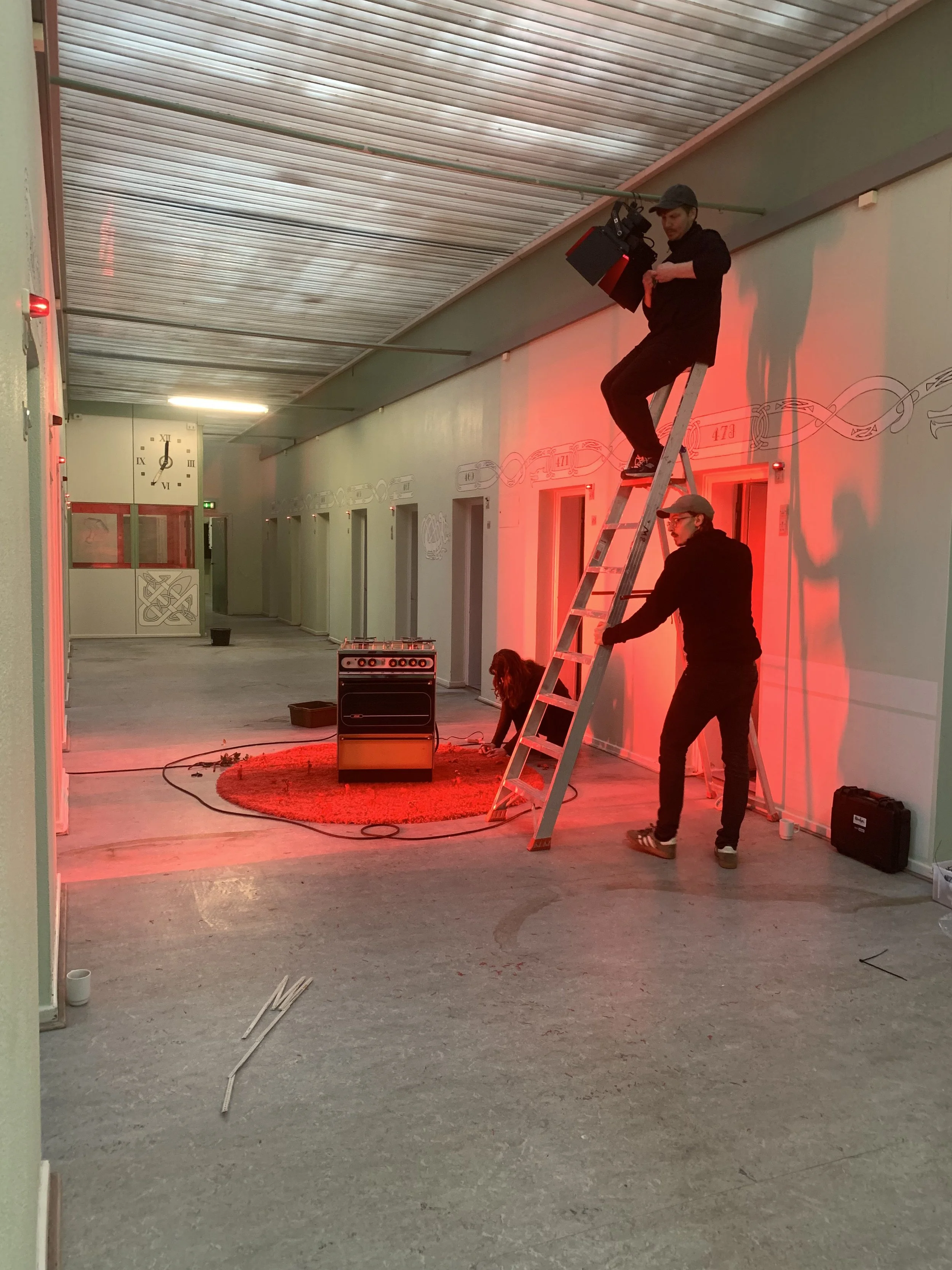 Tre personer arbejder i en gang med rødt lys. En person står på en stige og holder et filmlys, en anden står ved stigen | Dokumentarfilm | Retrograd | Esben Persson & Anders Mielcke | Filmproduktion Aarhus | GESTALT