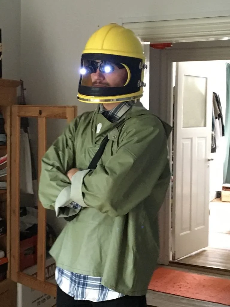 Person i grøn jakke med en gul hjelm, der har lys på fronten, står indendørs med armene krydset | Kortfilm | In Medias Ræs | Esben Persson | Filmproduktion Aarhus | GESTALT