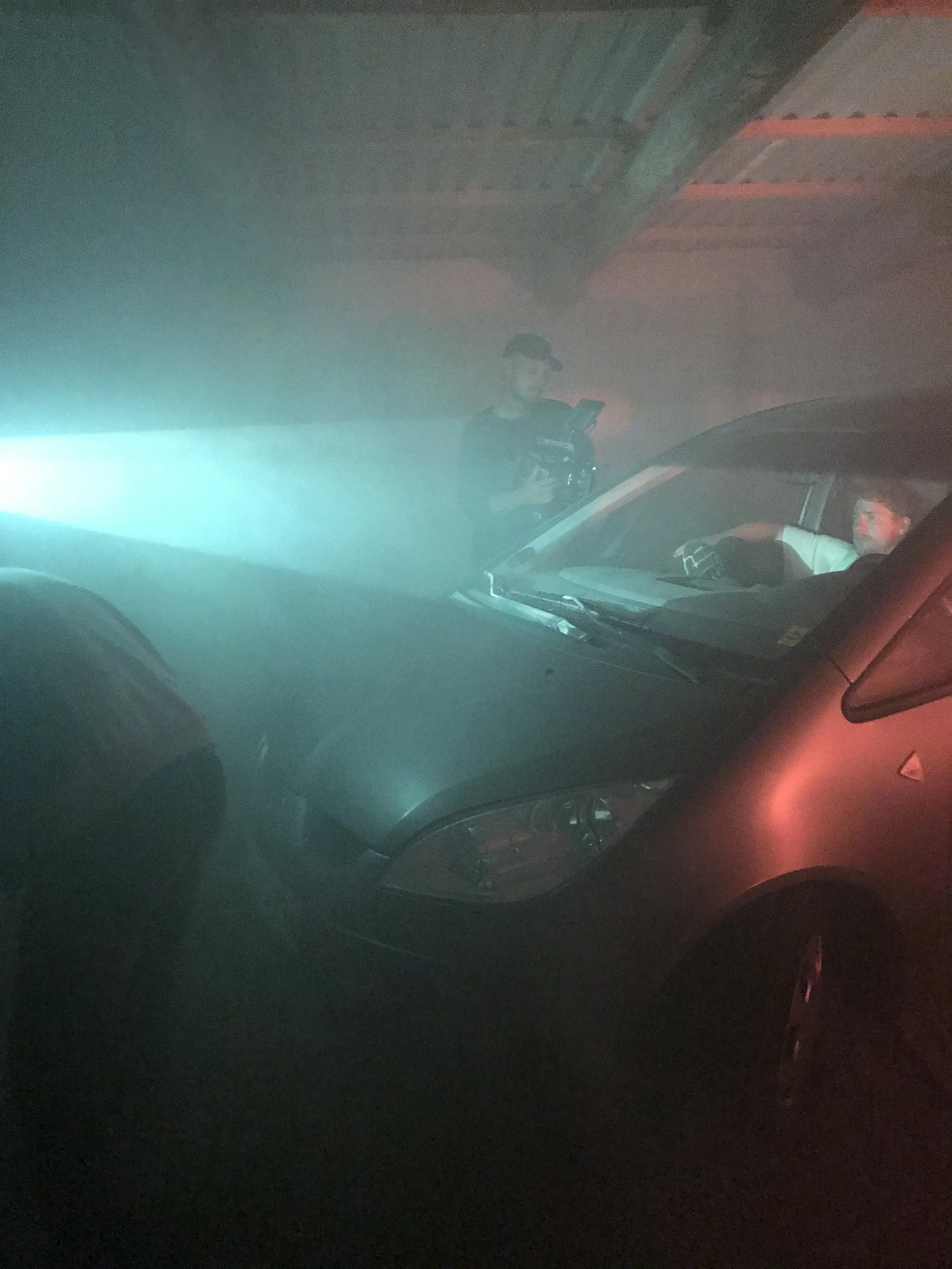 To personer i en bil i en dunkel garage med tåget lys, en person sidder foran i bilen og en anden står udenfor med et kamera og filmer | Kortfilm | Wild Wild Vesløs | Esben Persson | Filmproduktion Aarhus | GESTALT