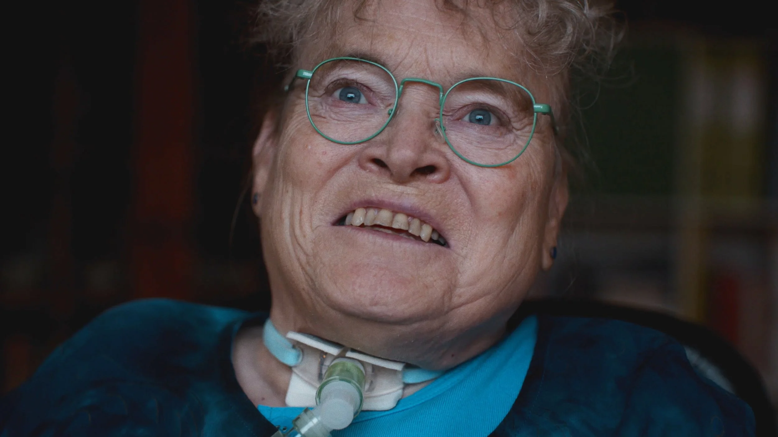 En ældre kvinde med briller og en tracheostomi i halsen, der smiler | Dokumentarfilm | Stemmer fra sær- og åndssvageforsorgen | Anders Mielcke Grønbæk | GESTALT