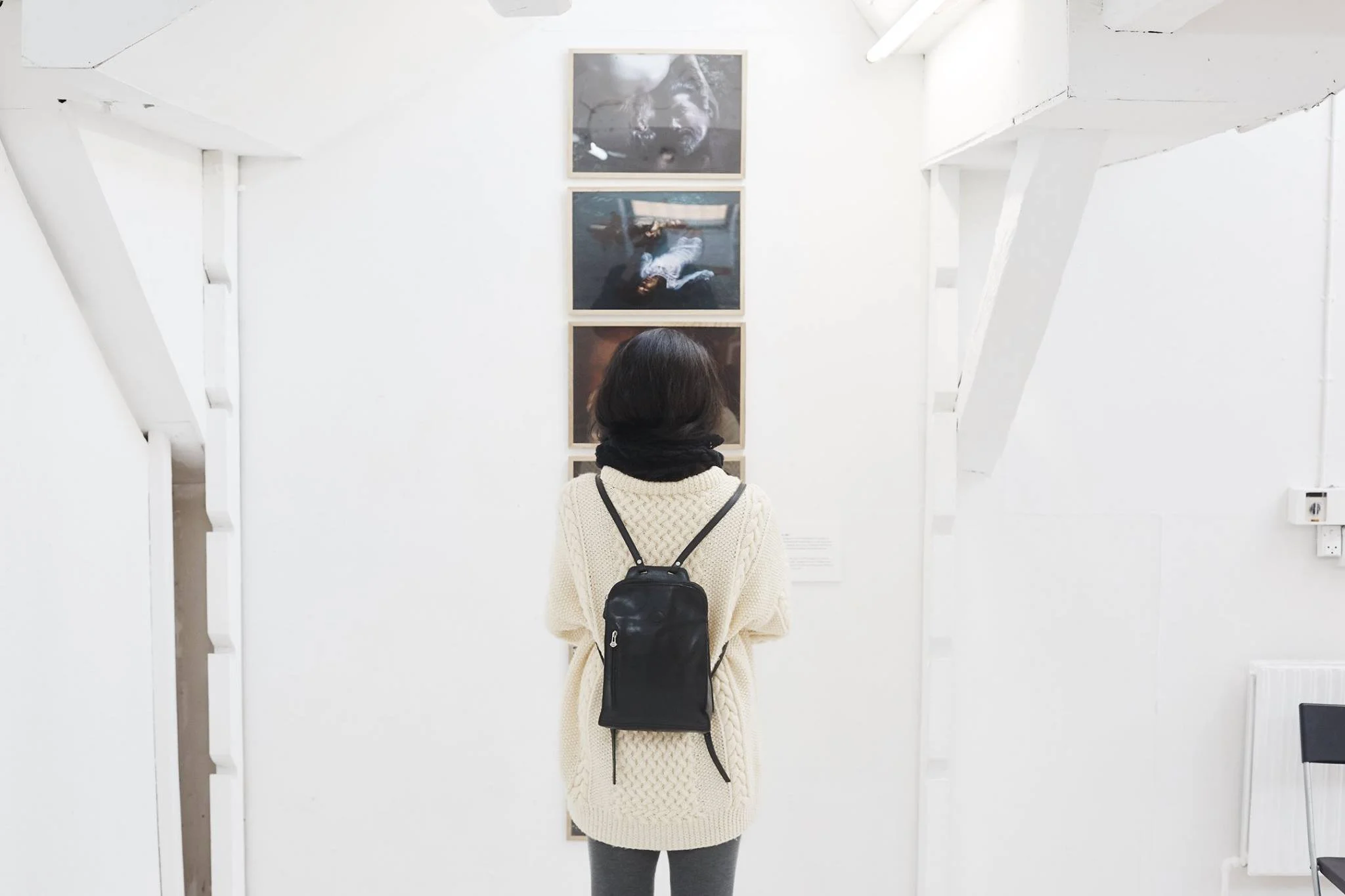 En person med mørkt hår, iført en beige strikket sweater og en sort rygsæk, står foran en væg med tre fotografier i en gallerikontekst | Aarhus Independent Pixels på Aarhus Kunstakademi | Eventproduktion | Filmproduktion Aarhus | GESTALT