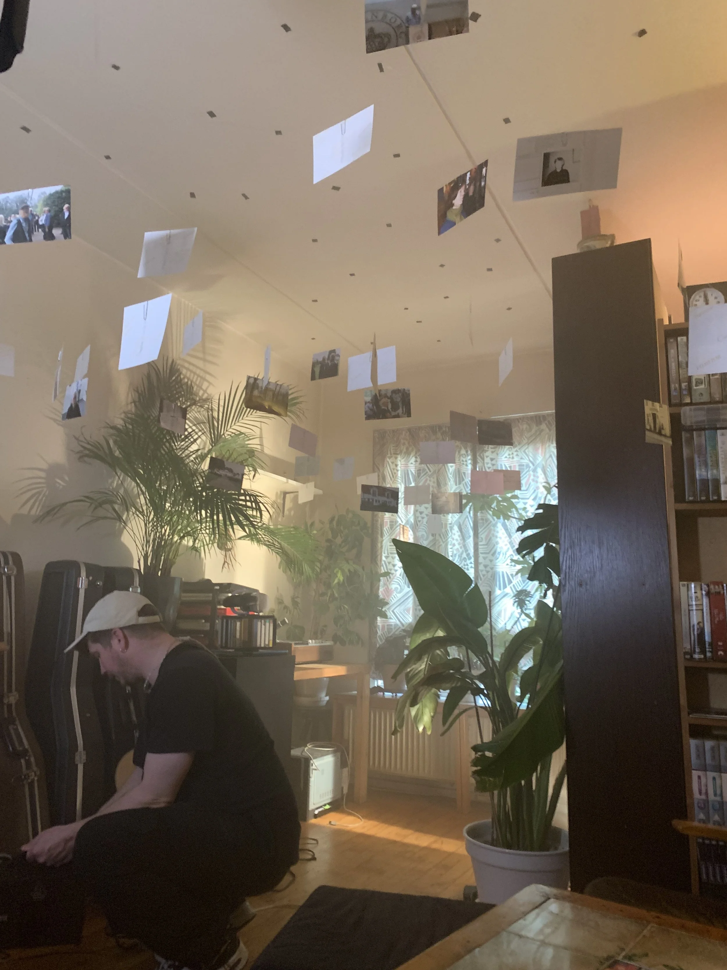 Et indendørs rum med grønne planter, et møbel, en person med kasket, og en loftdekoration med hængende billeder | Dokumentarfilm | Retrograd | Esben Persson & Anders Mielcke | Filmproduktion Aarhus | GESTALT