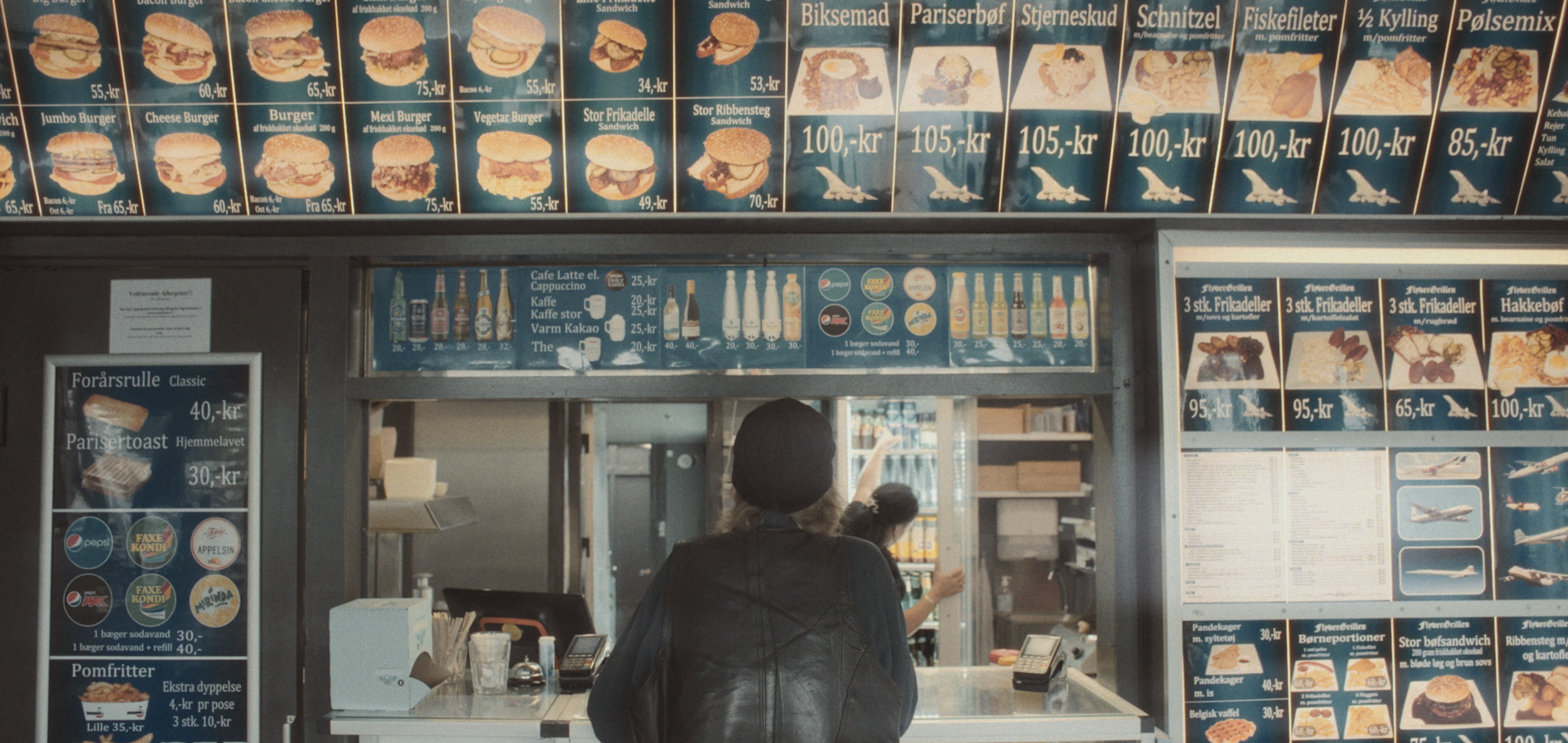 Billede af en take-away-menu på en grill der viser forskellige burger og sandwich-muligheder med priser. Der er en person foran kassen | Dokumentarfilm | Retrograd | Esben Persson & Anders Mielcke | Filmproduktion Aarhus | GESTALT