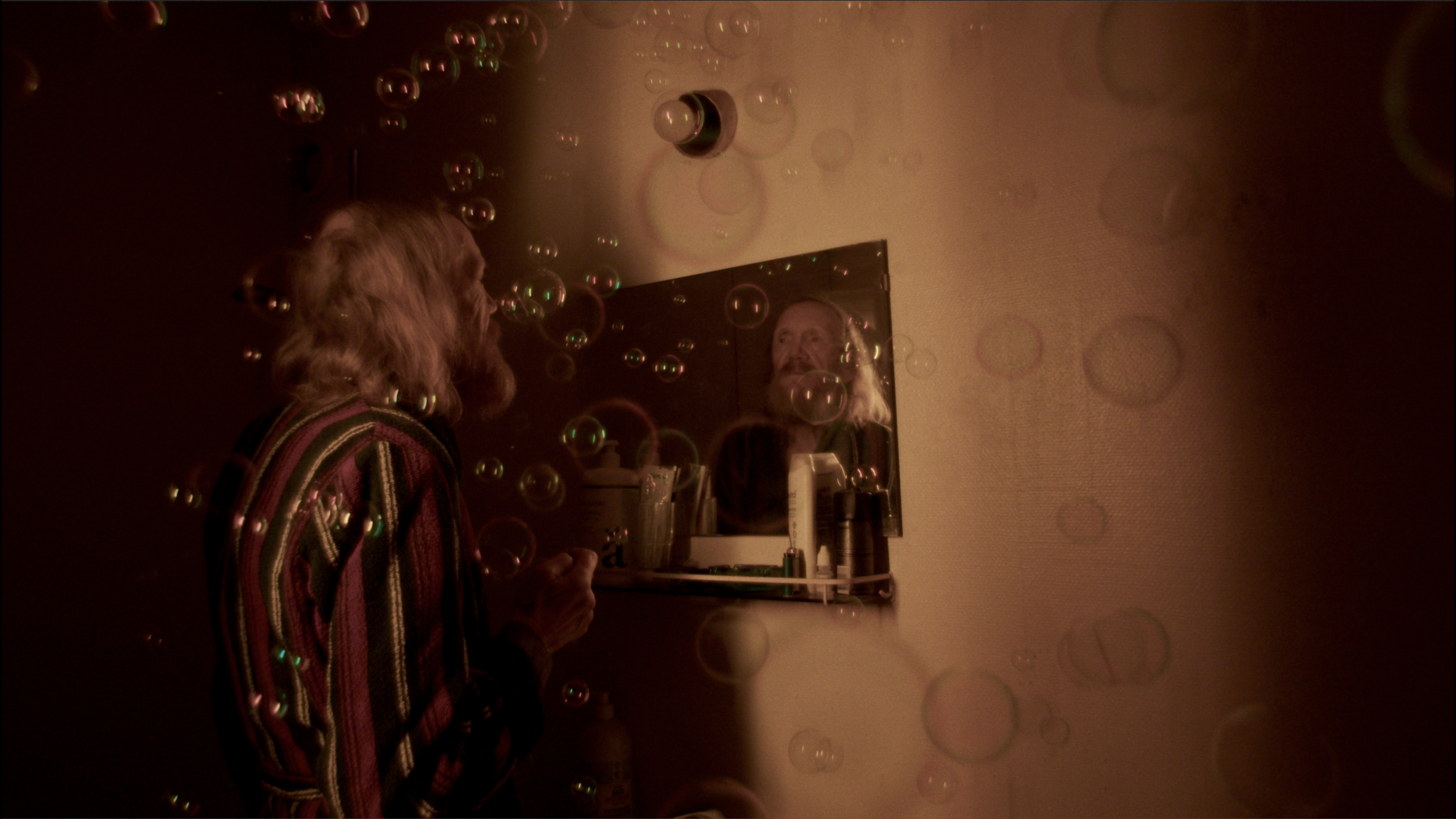 En ældre person med langt, lyst hår og lyst skæg kigger i et spejl over vasken. Der er mange såpabobler i rummet, og billedet har en dæmpet belysning | Dokumentarfilm | Retrograd | Esben Persson & Anders Mielcke | Filmproduktion Aarhus | GESTALT