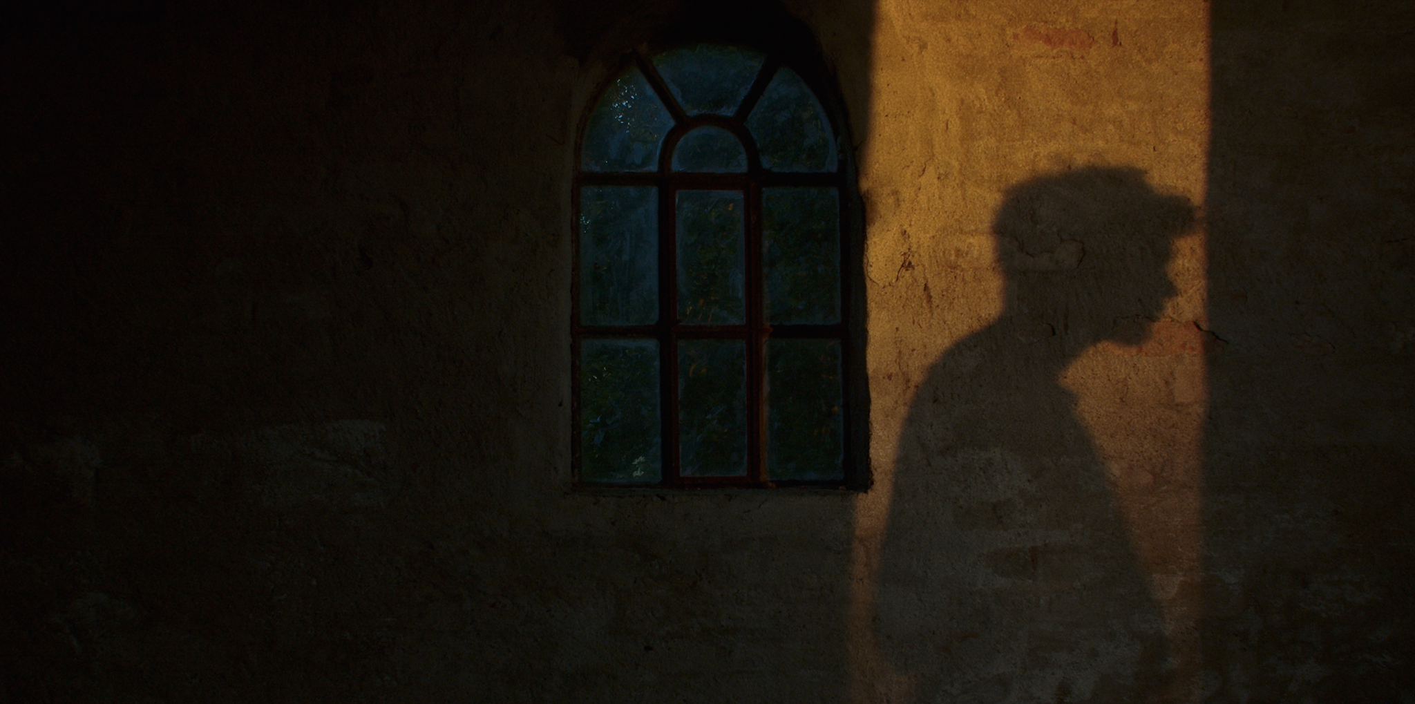 En skygge af en person med krøllet hår, der står foran et gammelt stenvindue, med skyggen der falder på en væg ved solnedgang | Kortfilm | Wild Wild Vesløs | Esben Persson | Filmproduktion Aarhus | GESTALT
