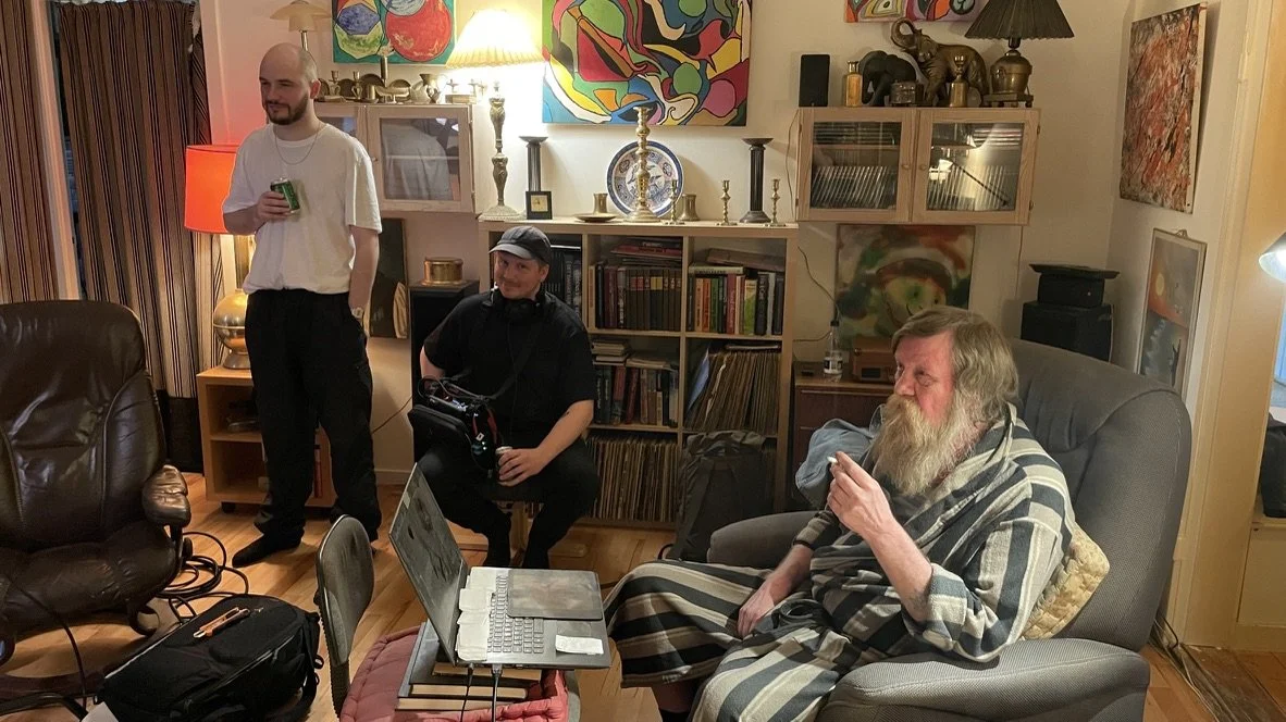 Tre personer i et stueopstilling med bøger, lamper og kunst på væggen, to sidder og én står med en øl, mens den tredje bruger en bærbar computer | Dokumentarfilm | Retrograd | Esben Persson & Anders Mielcke  | Filmproduktion Aarhus | GESTALT