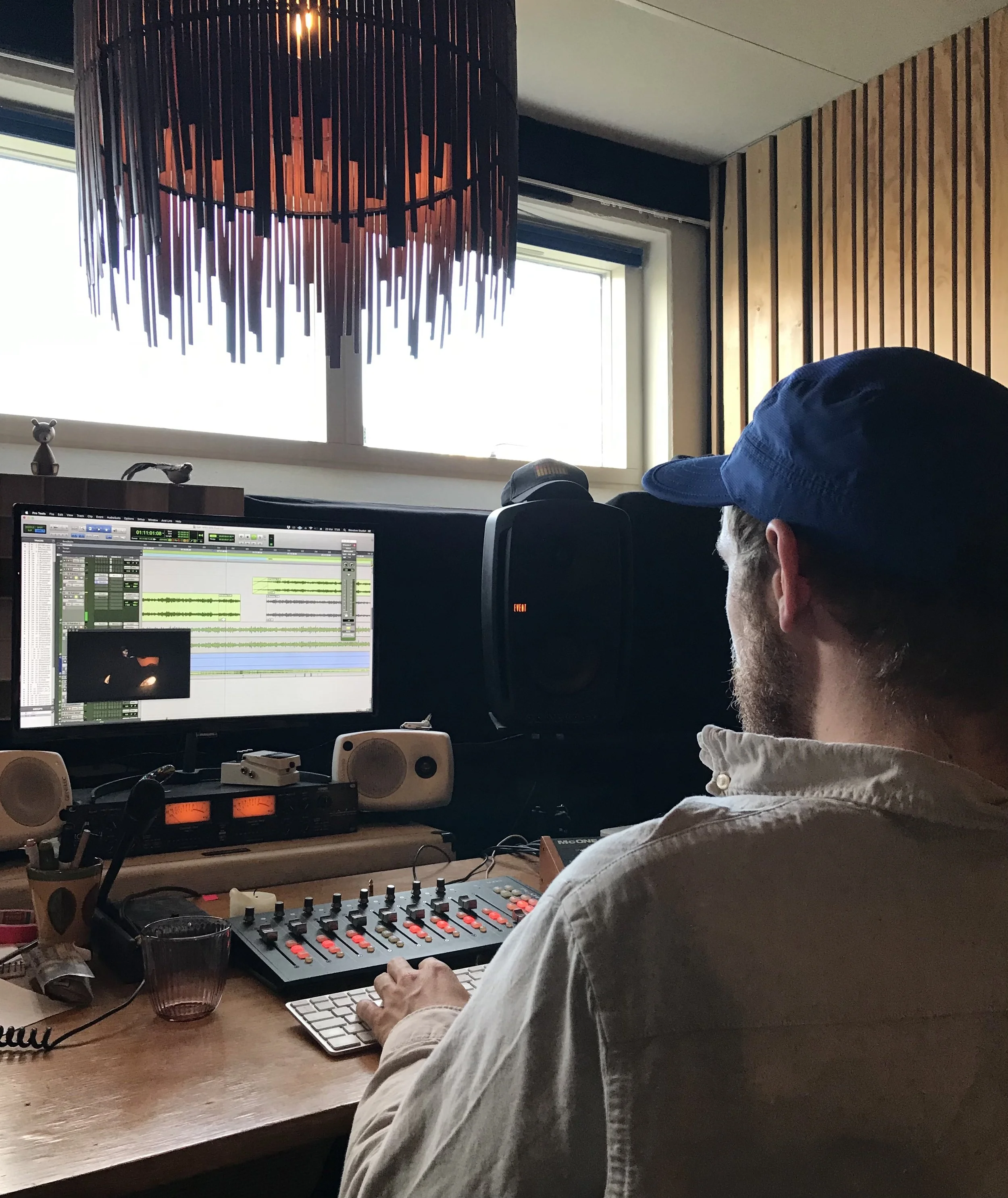 Person arbejder i et lydstudie med computer, mixer og højttalere, med en stor lampe hængende over arbejdspladsen | Musikkomposition | Lyddesign | Filmproduktion Aarhus | GESTALT