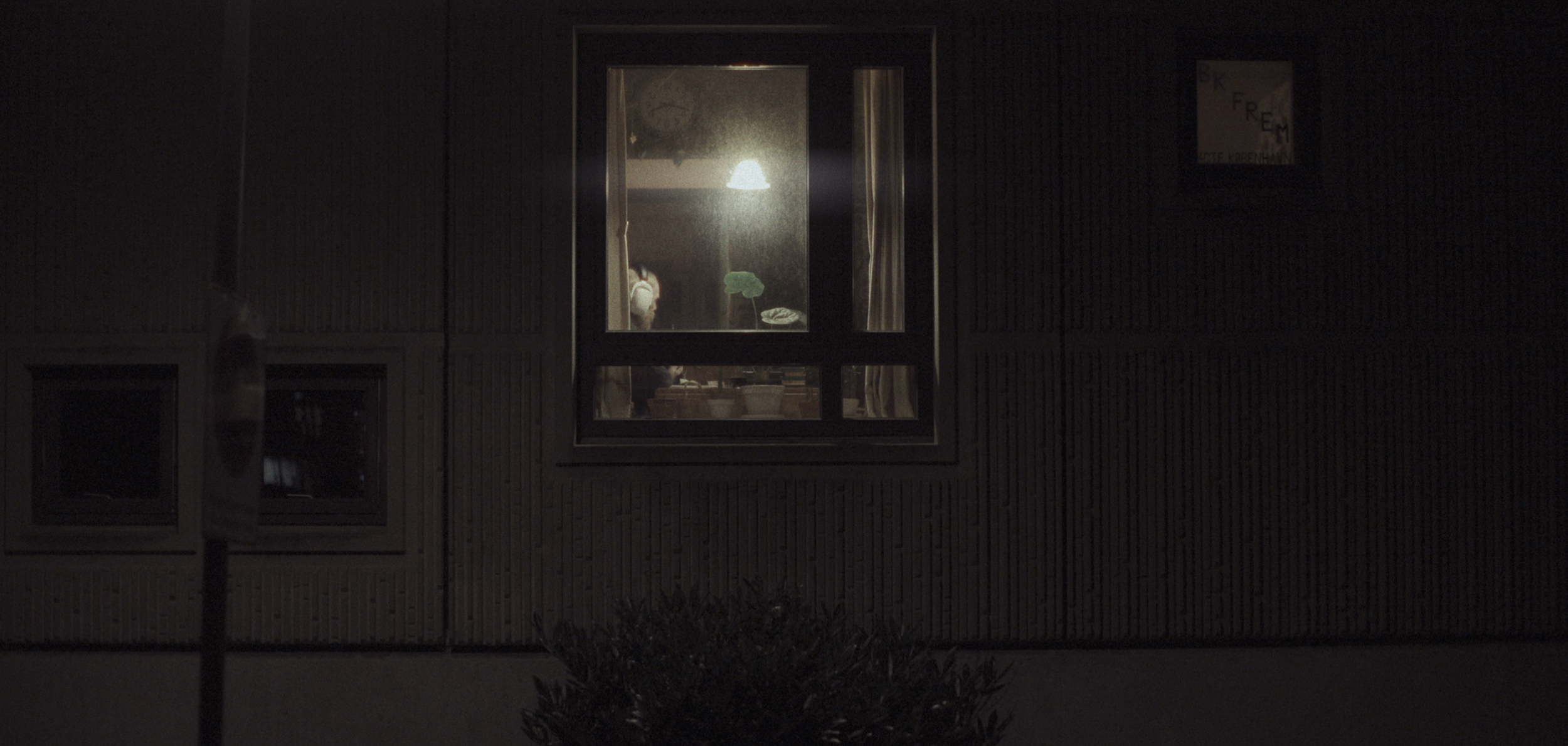 Et motiv taget udenfor i natten, der viser en bygning med en oplyst vinduesramme, hvor en person sidder ved et bord indendørs. Han sidder ved en båndoptager | Dokumentarfilm | Retrograd | Esben Persson & Anders Mielcke | Filmproduktion Aarhus | GESTA