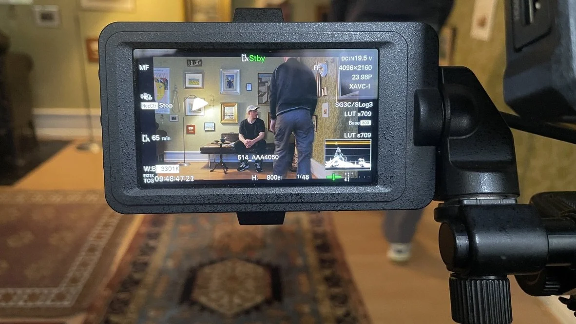 En filmkamera, der filmer en person, der sidder på en stol i en stue med vægge dekoreret med billeder | Dokumentarfilm | Retrograd | Esben Persson & Anders Mielcke  | Filmproduktion Aarhus | GESTALT