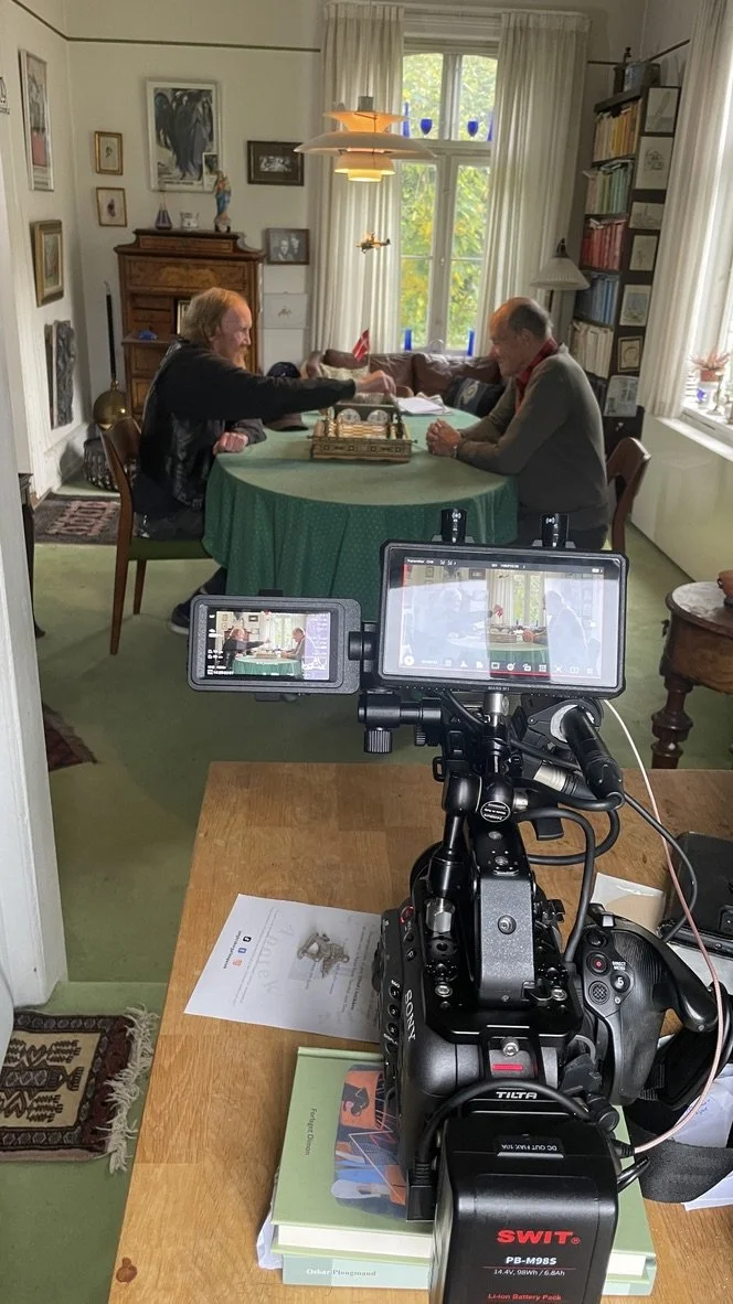 To ved et bord, der spiller skak i et lyst værelse med store vinduer, lange hvide gardiner, billeder på væggen, og bøger på en reol | Dokumentarfilm | Retrograd | Esben Persson & Anders Mielcke  | Filmproduktion Aarhus | GESTALT