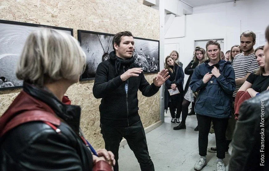 En gruppe mennesker i et fotogalleri. Han gestikulerer med hænderne, mens de andre lytter opmærksomt. mænd holder oplæg | Film- og fotofestival på Aarhus Kunstakademi ved navn Aarhus Independent Pixels | Eventbilleder | GESTALT